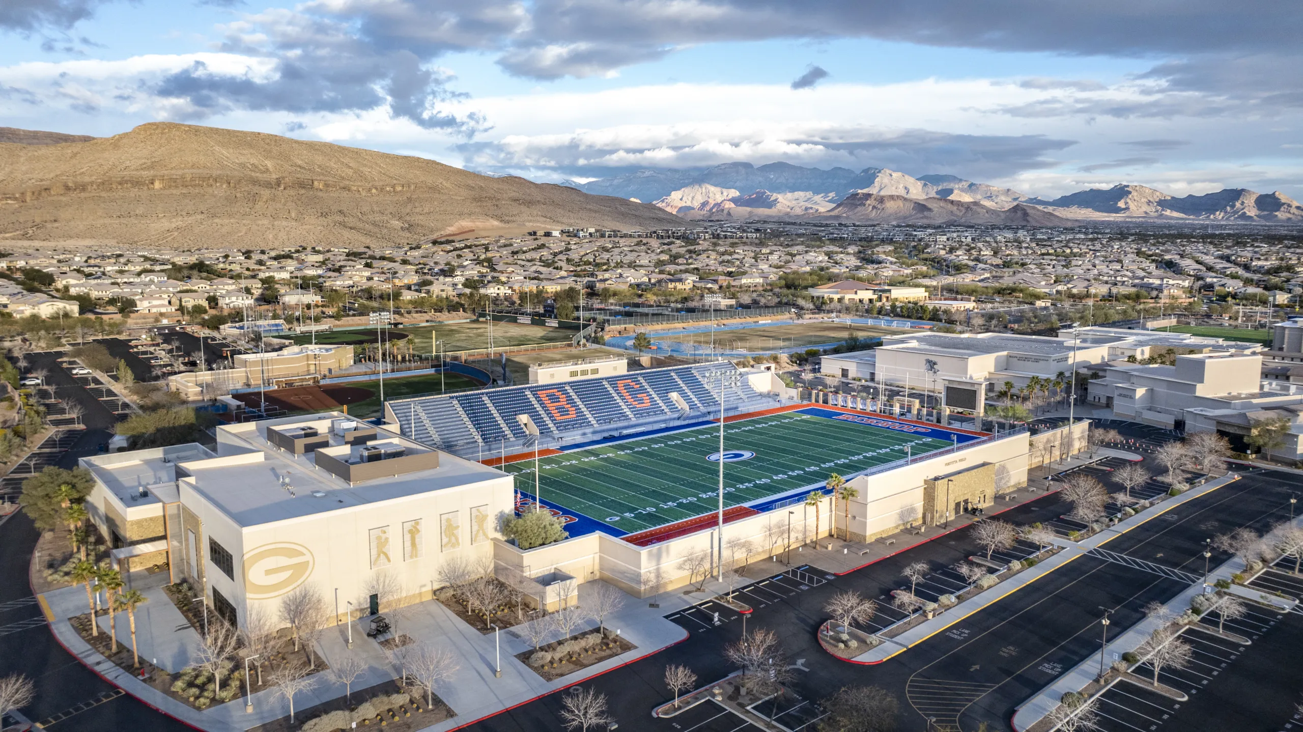 Aerial view of a large high school football stadium and campus, with mountains and a residential neighborhood in the background under a partly cloudy sky.