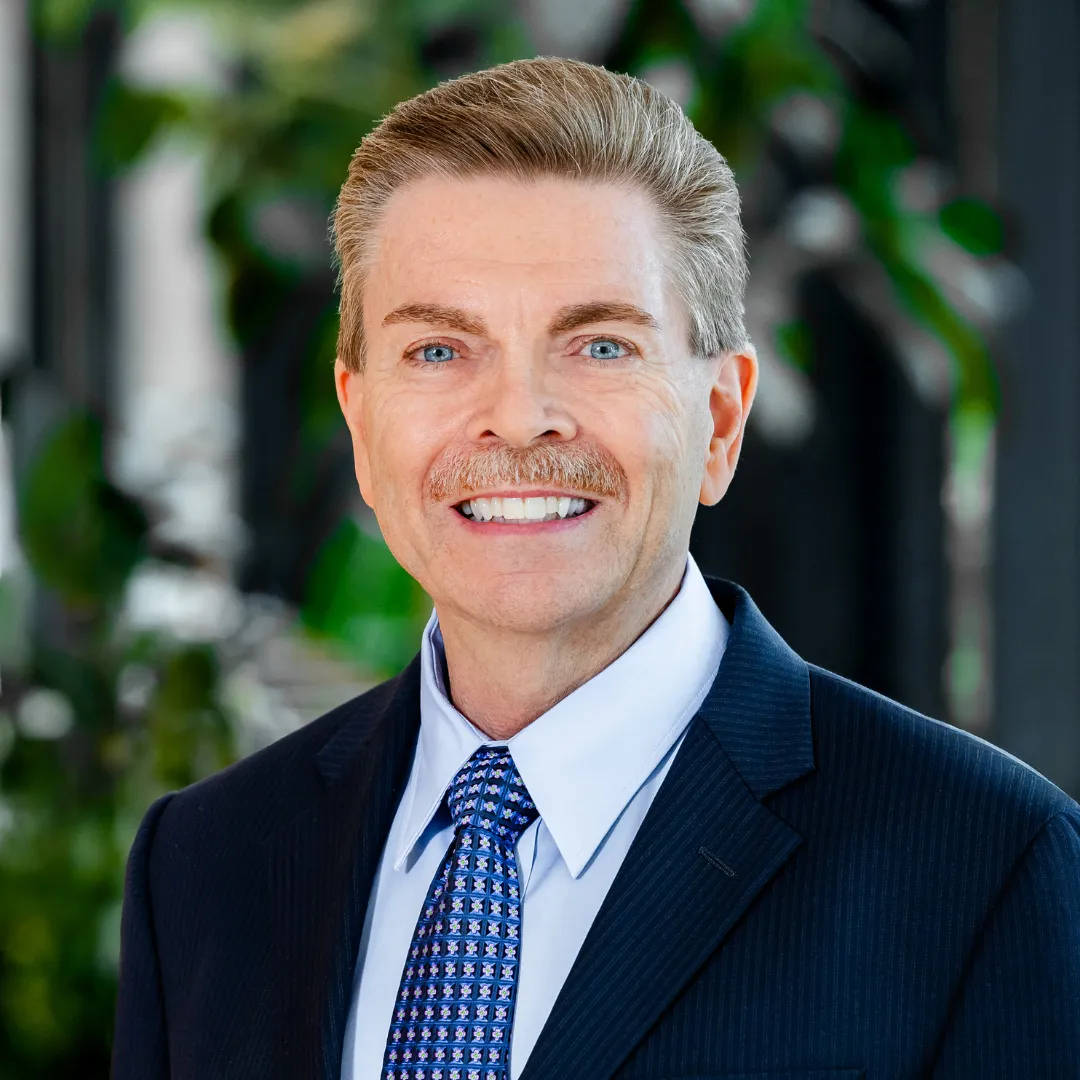 Brian Headshot Smiling man with light brown hair, mustache, and blue eyes, wearing a dark suit, blue patterned tie, and white shirt. Green foliage and a window are blurred in the background.