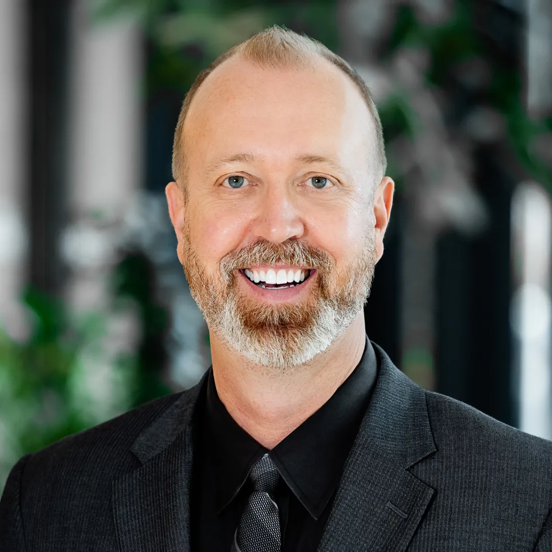 Chad Headshot A smiling man with short blonde hair and a trimmed beard, wearing a dark suit, shirt, and tie, stands in front of a blurred background with green plants.
