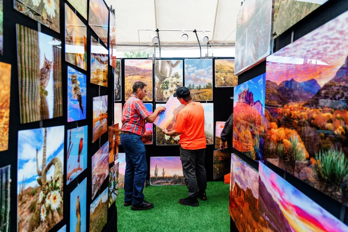 Two people stand in a booth displaying vibrant landscape photos. They are talking and pointing at images of deserts, mountains, and cacti under a white tent on a green, grassy floor.
