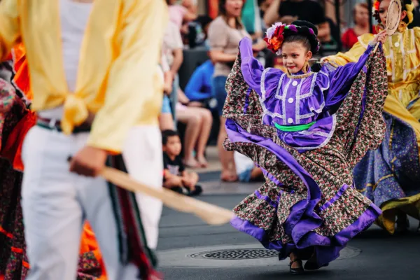 A young girl in a vibrant purple traditional dress dances joyfully in a street parade, surrounded by other performers and onlookers.