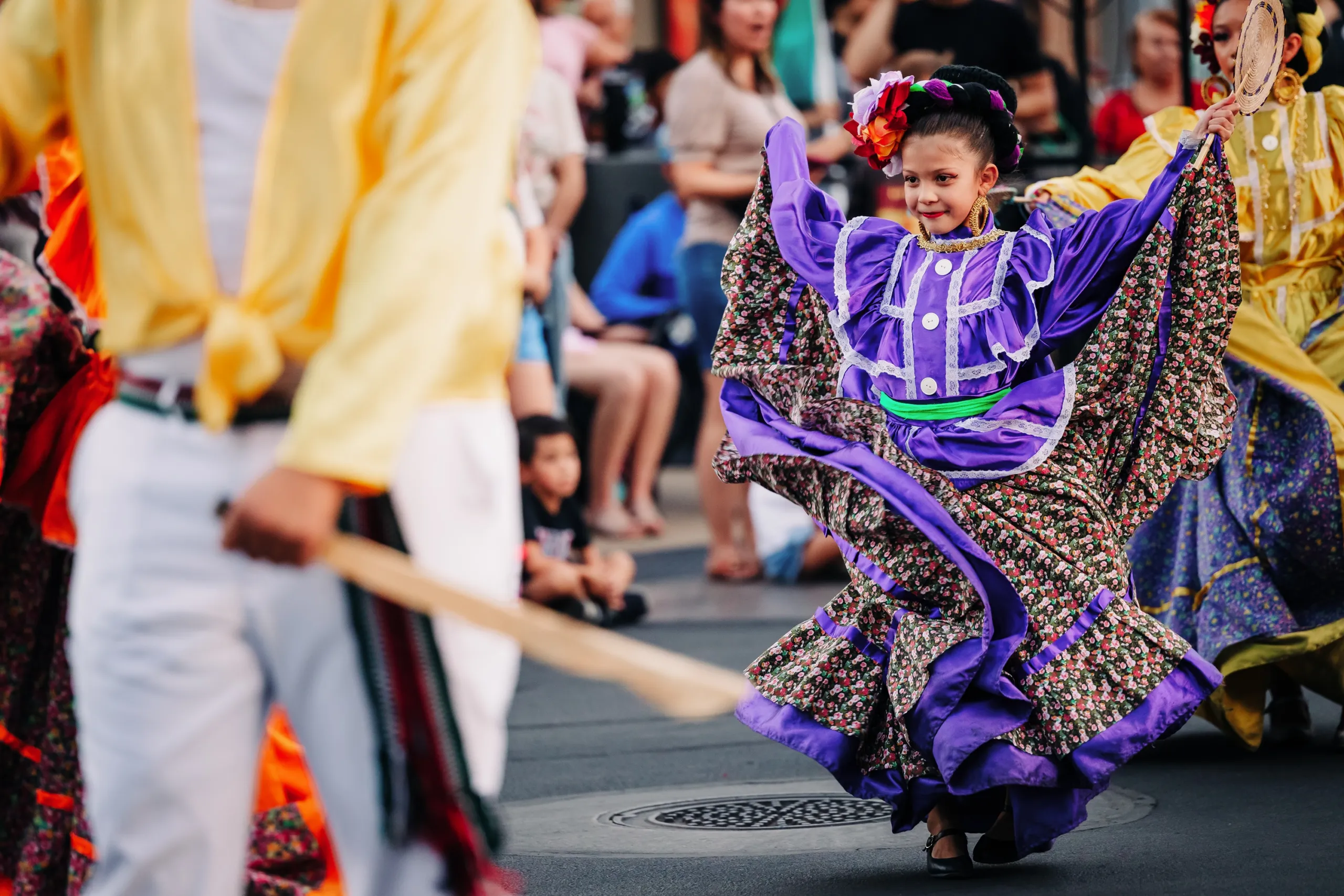 A young girl in a vibrant purple traditional dress dances joyfully in a street parade, surrounded by other performers and onlookers.