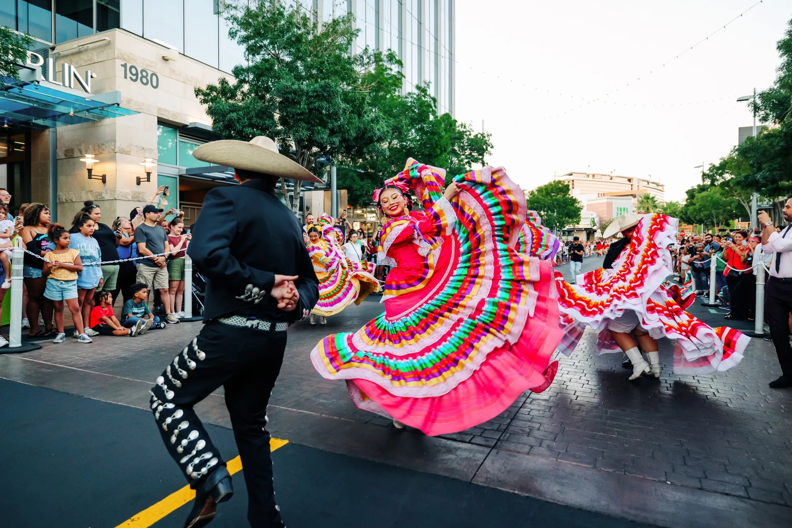 Dancers in colorful traditional Mexican attire perform folkloric dance on a city street, surrounded by a crowd of onlookers.