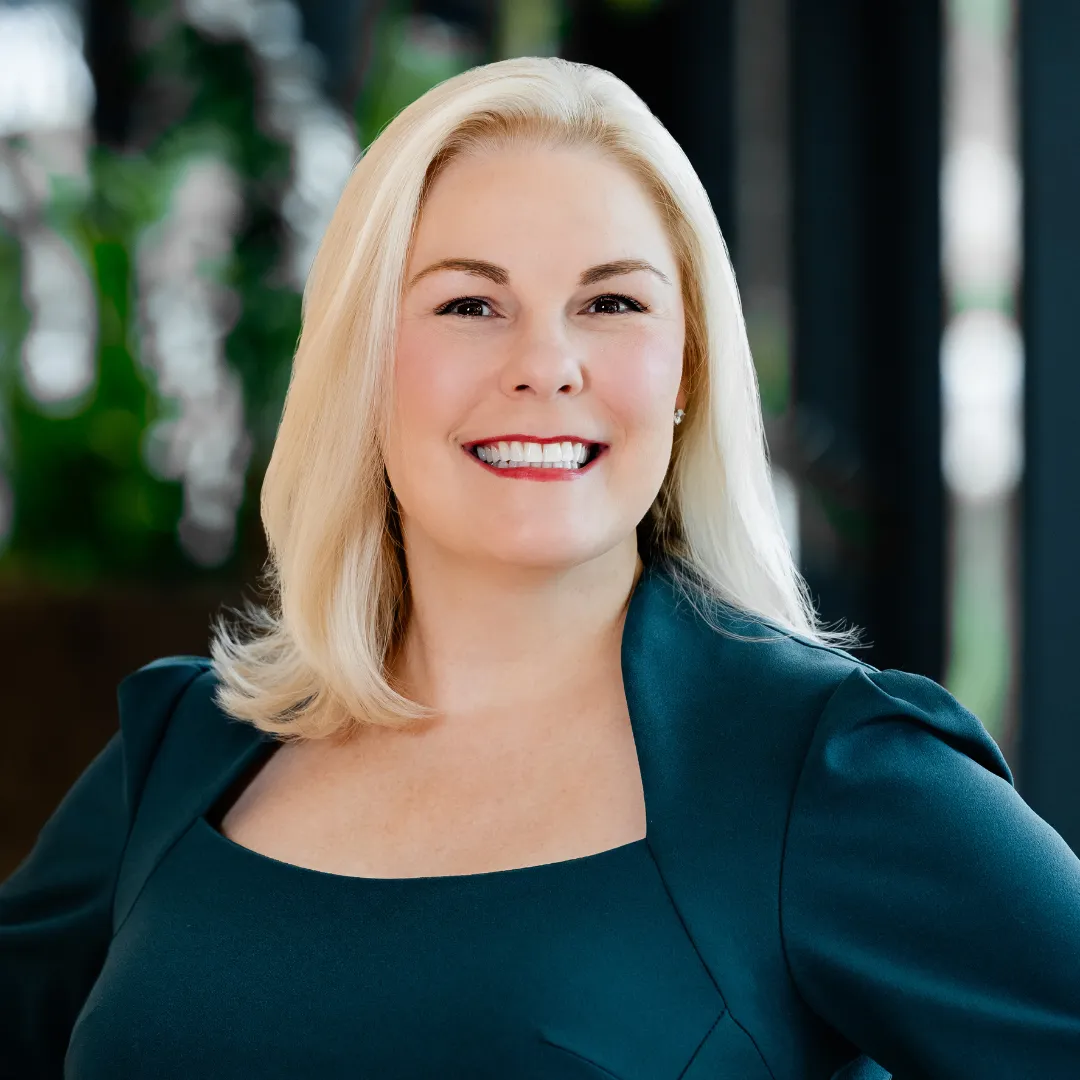 Danielle Headshot A woman with blonde hair, wearing a dark green top, smiles confidently while standing indoors with blurred greenery in the background.