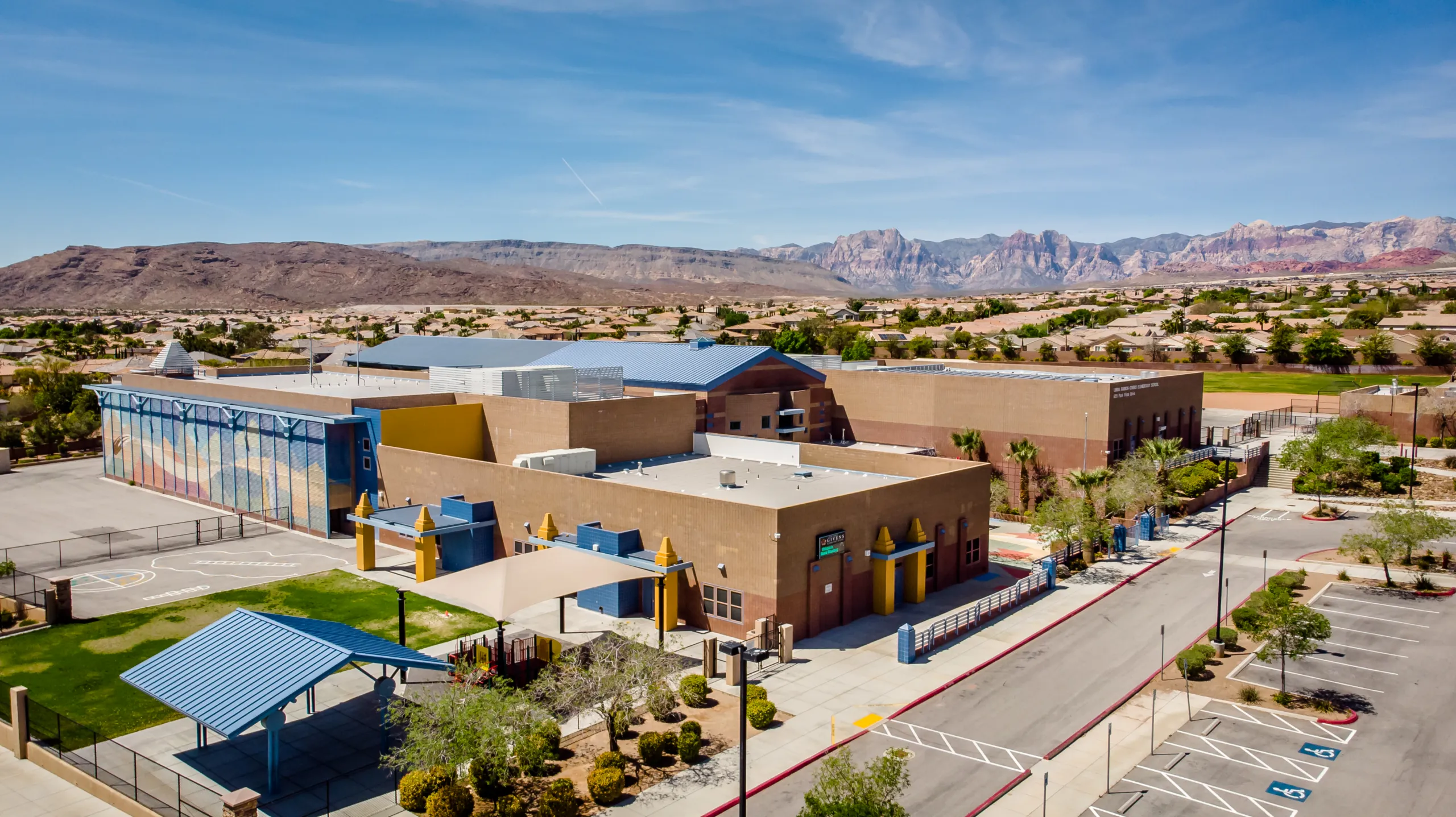 Aerial view of a modern school with tan and blue buildings, a playground, and surrounding desert landscape, with mountains in the background under a clear blue sky.