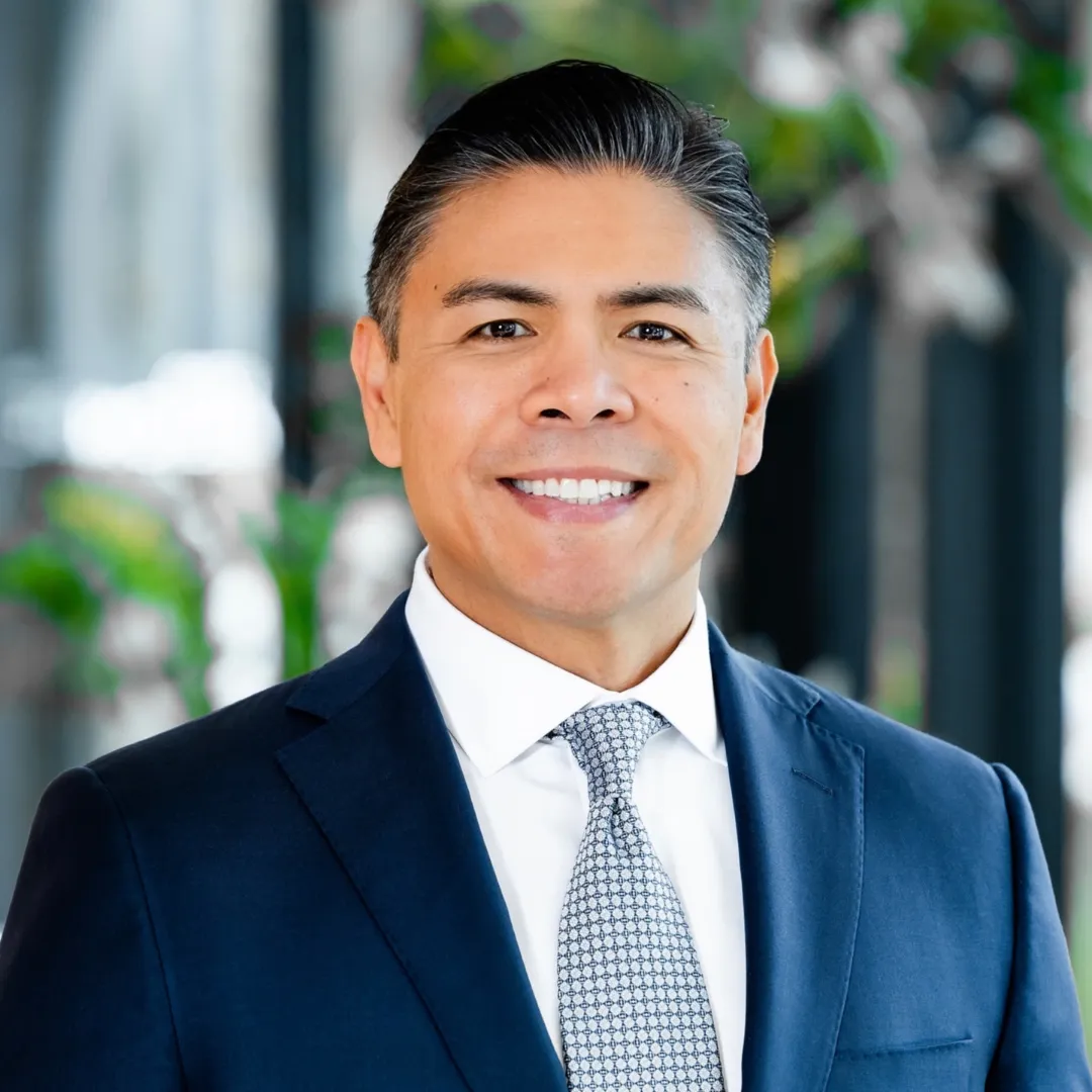 Jose Headshot Smiling man in a navy suit and patterned tie stands indoors, with blurred greenery and windows in the background.