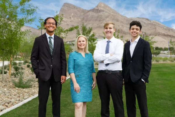 Four people in business attire stand smiling outdoors on grass with mountains and blue sky in the background. Two men wear suits, one wears a shirt and tie, and one woman wears a teal dress.
