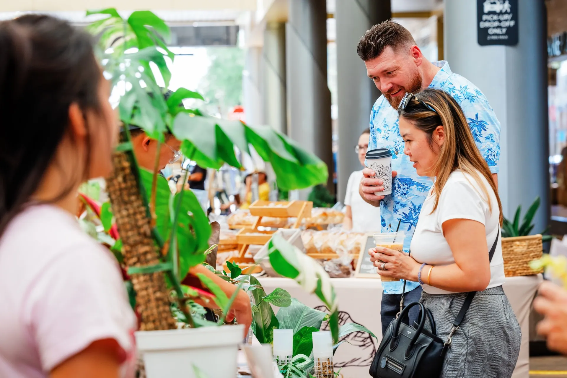 Sunday Stroll Two people holding drinks talk to a vendor at an outdoor market stall with plants and baked goods. Other customers and greenery are visible in the lively scene.