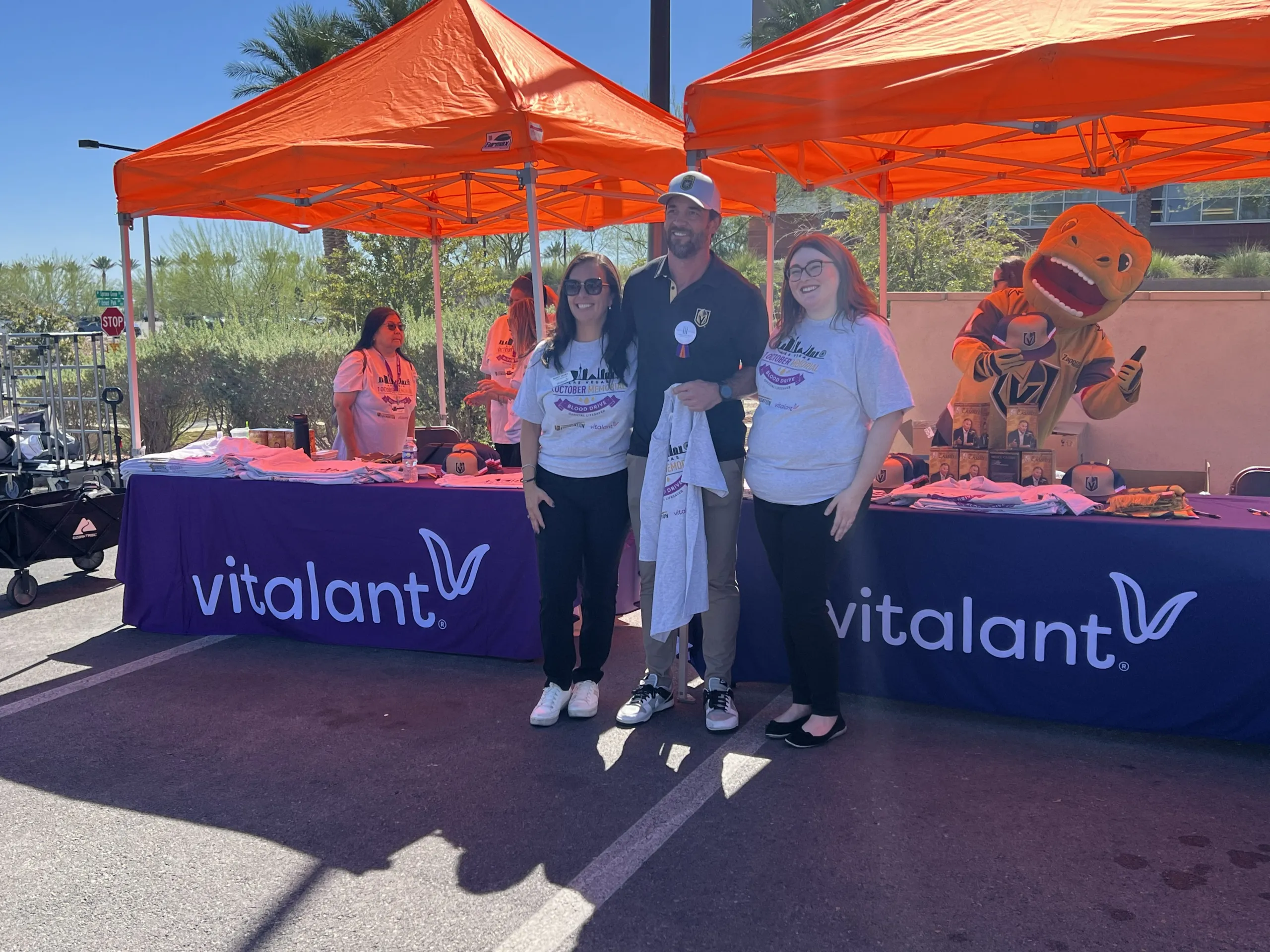 1 October Memorial Blood Drive 2025 05 Three people pose and smile in front of two orange tents with Vitalant table covers. A mascot in a yellow suit is on the right. The event appears to be outdoors with sunny weather and trees in the background.