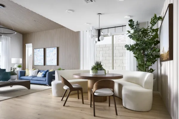 Modern living and dining area with a round wooden table, curved white bench, two wooden chairs, large indoor plant, light wood floors, blue sofa, and wall art. Room is bright with natural light from large windows.