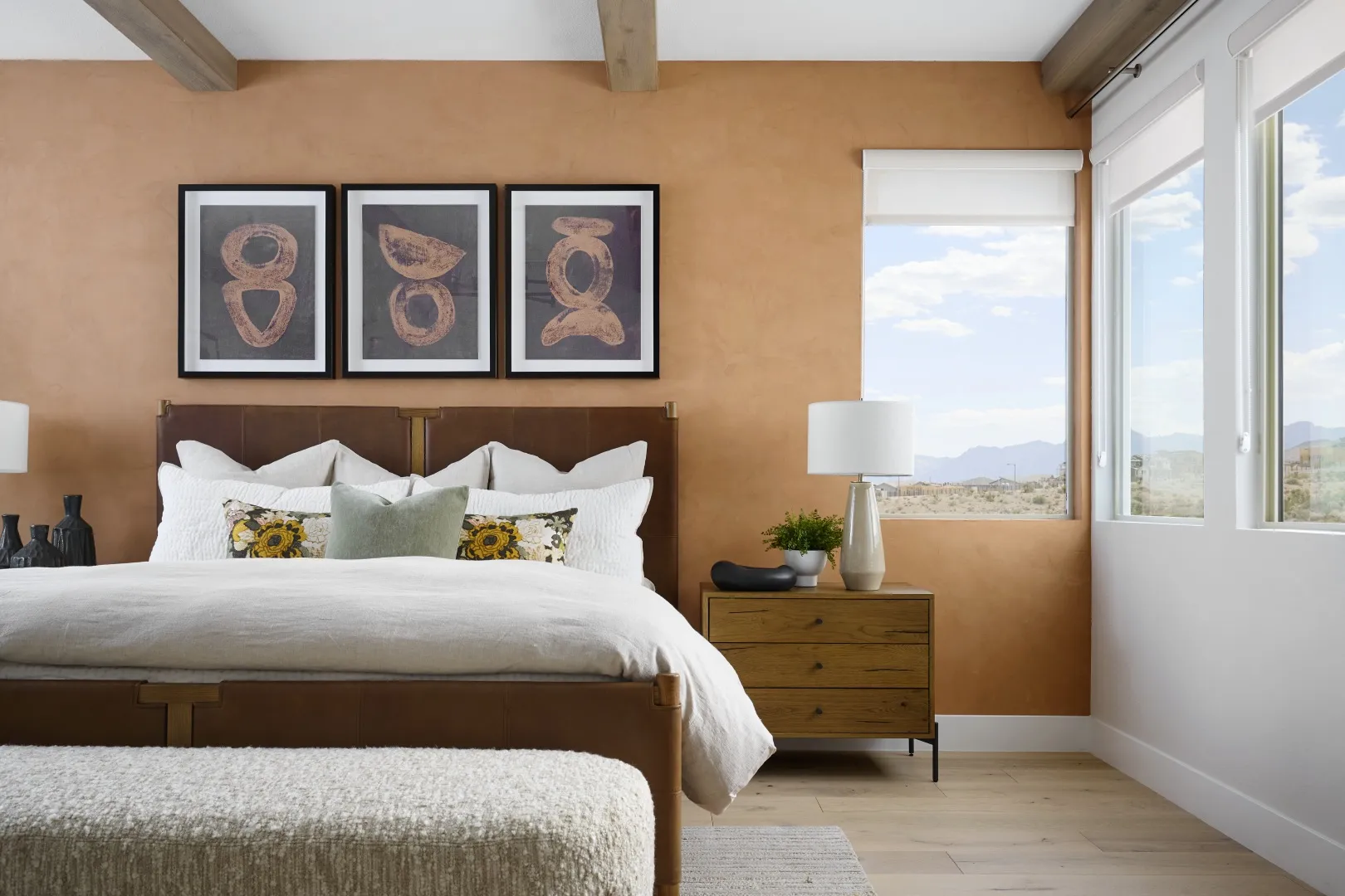 A modern bedroom with a brown accent wall, a bed with white bedding and decorative pillows, three abstract artworks above the headboard, a wooden nightstand, and large windows showing a desert landscape.