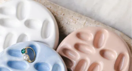 Artist Directory A gold ring with a green and a clear stone rests on a blue ceramic dish, next to white and pink dishes with indented designs, all placed on a textured surface.