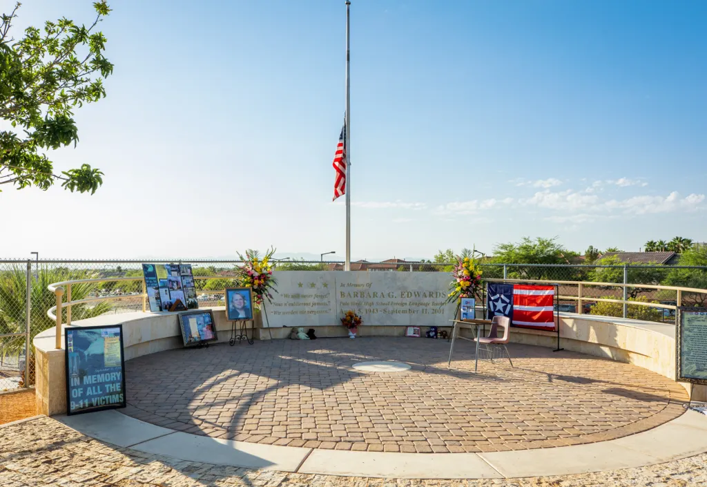 A memorial with an American flag at half-staff, flowers, photos, and tribute items displayed in a circular area under a clear sky, honoring victims.