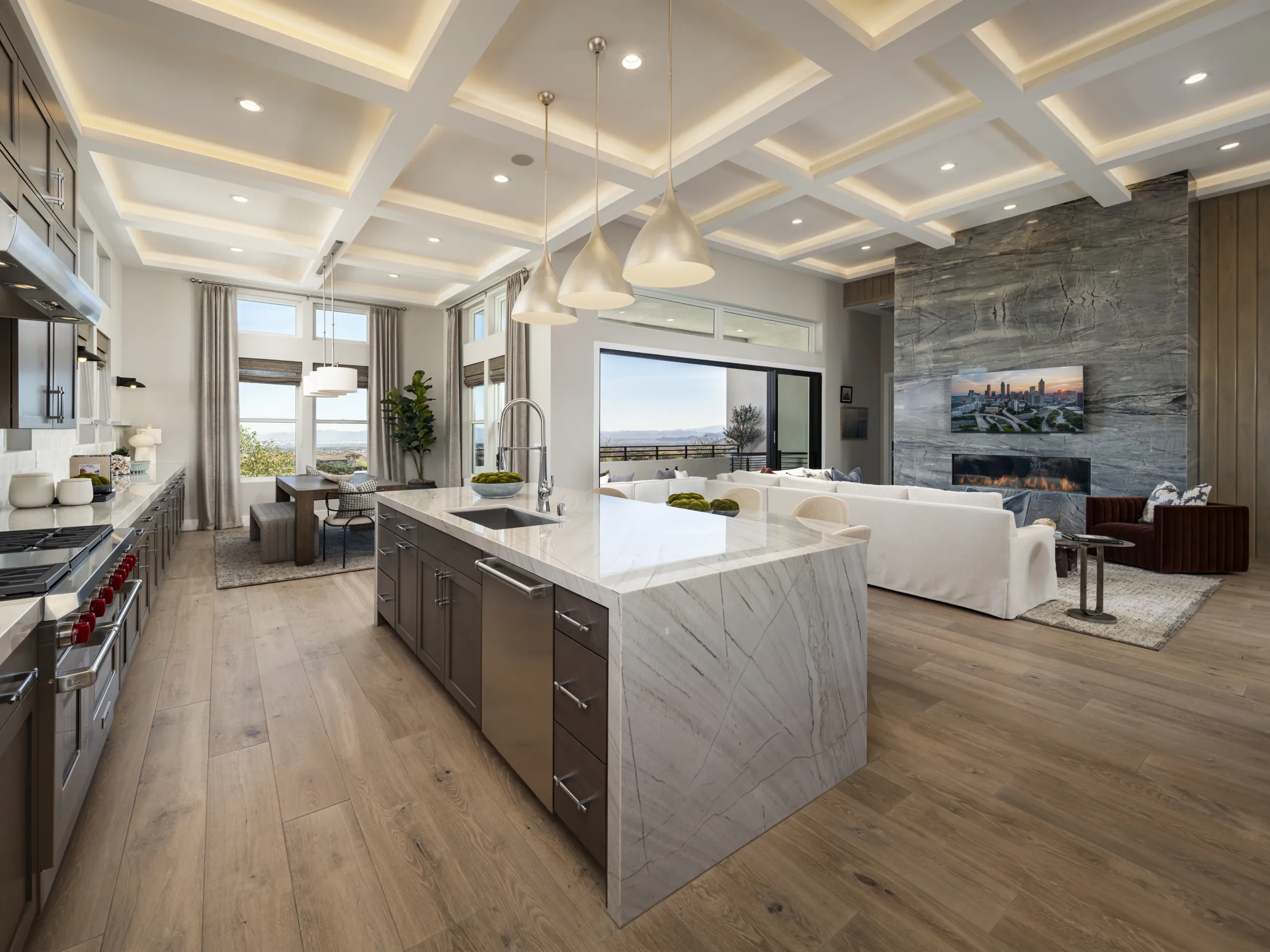 Modern open-concept kitchen and living room with a marble island, pendant lights, wood floors, large windows, a fireplace, and a TV above it. Ocean view visible through the windows.