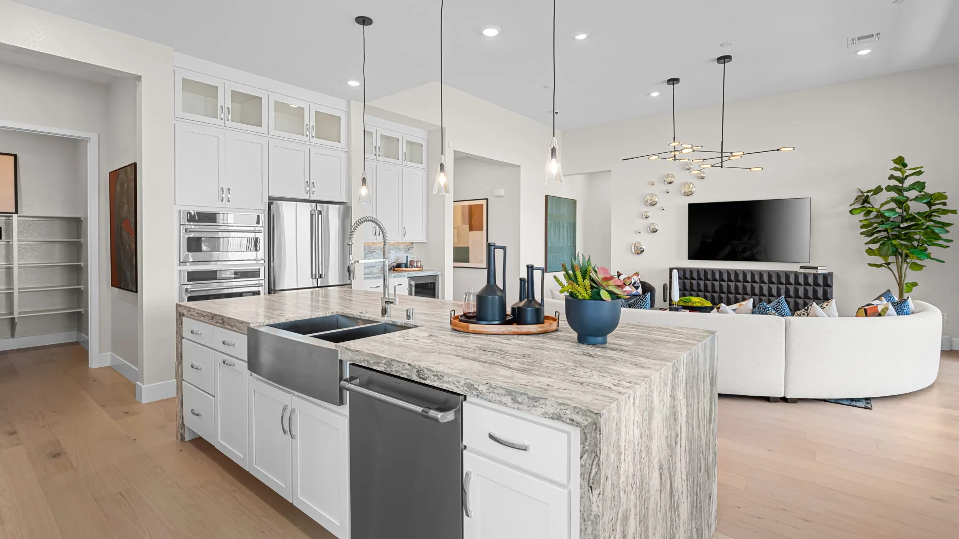 Modern kitchen with a large island, white cabinets, stainless steel appliances, pendant lights, and an open living area with a TV, round sofa, and decorative plants in a bright, spacious room.