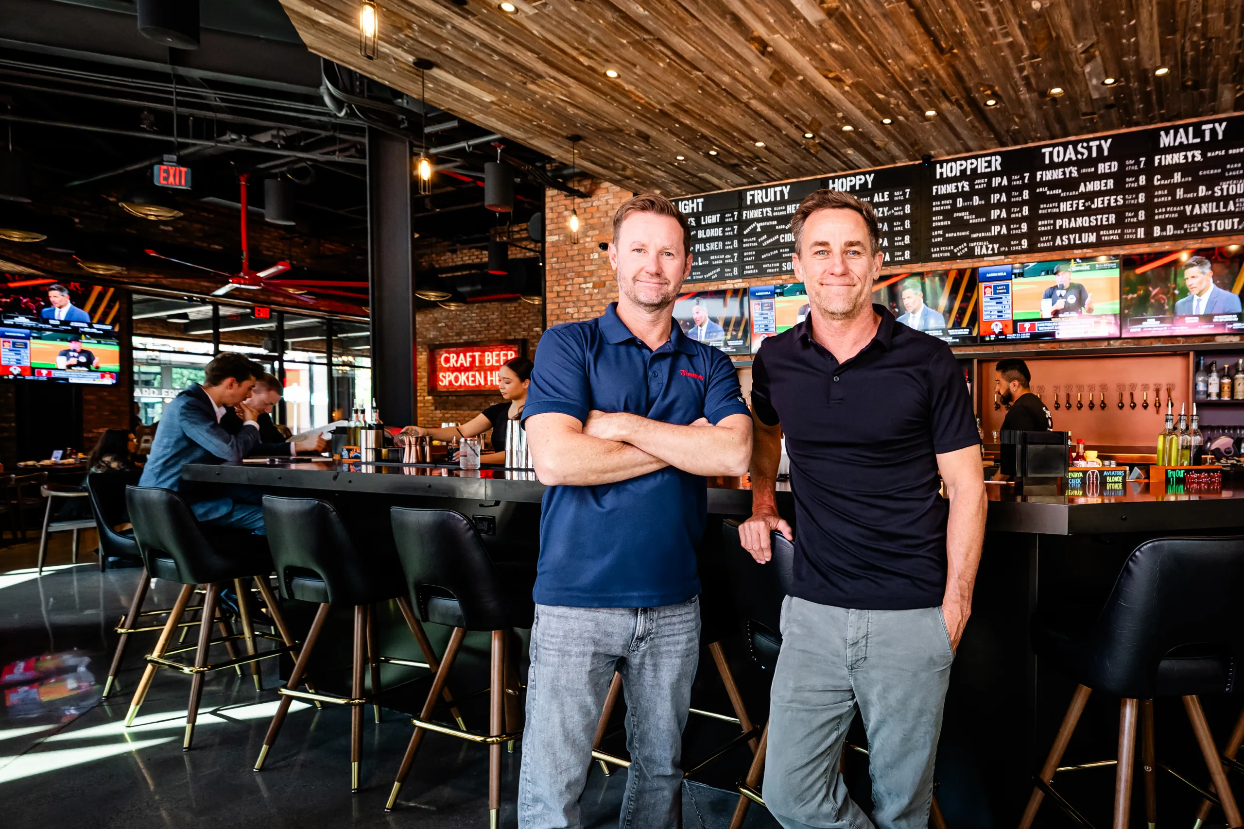 Two men stand smiling in front of a bar with TVs and neon signs. Other patrons sit at the bar, and the restaurant has a modern wood and metal design.