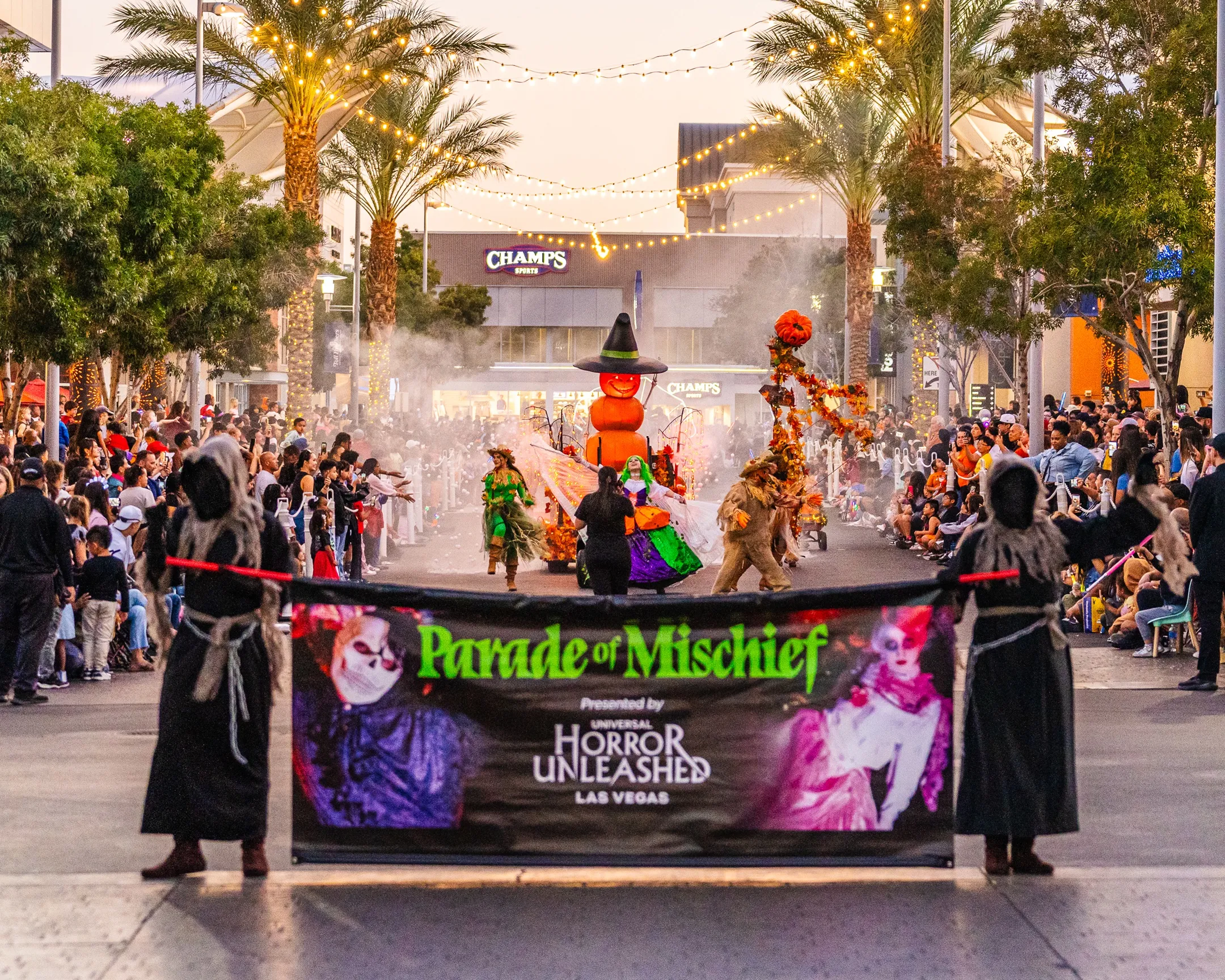 DTSHalloweenParadeExtras-28 People in costumes hold a Parade of Mischief banner on a lively, decorated street. A large witch balloon floats behind them, as spectators watch amid palm trees and glowing lights.