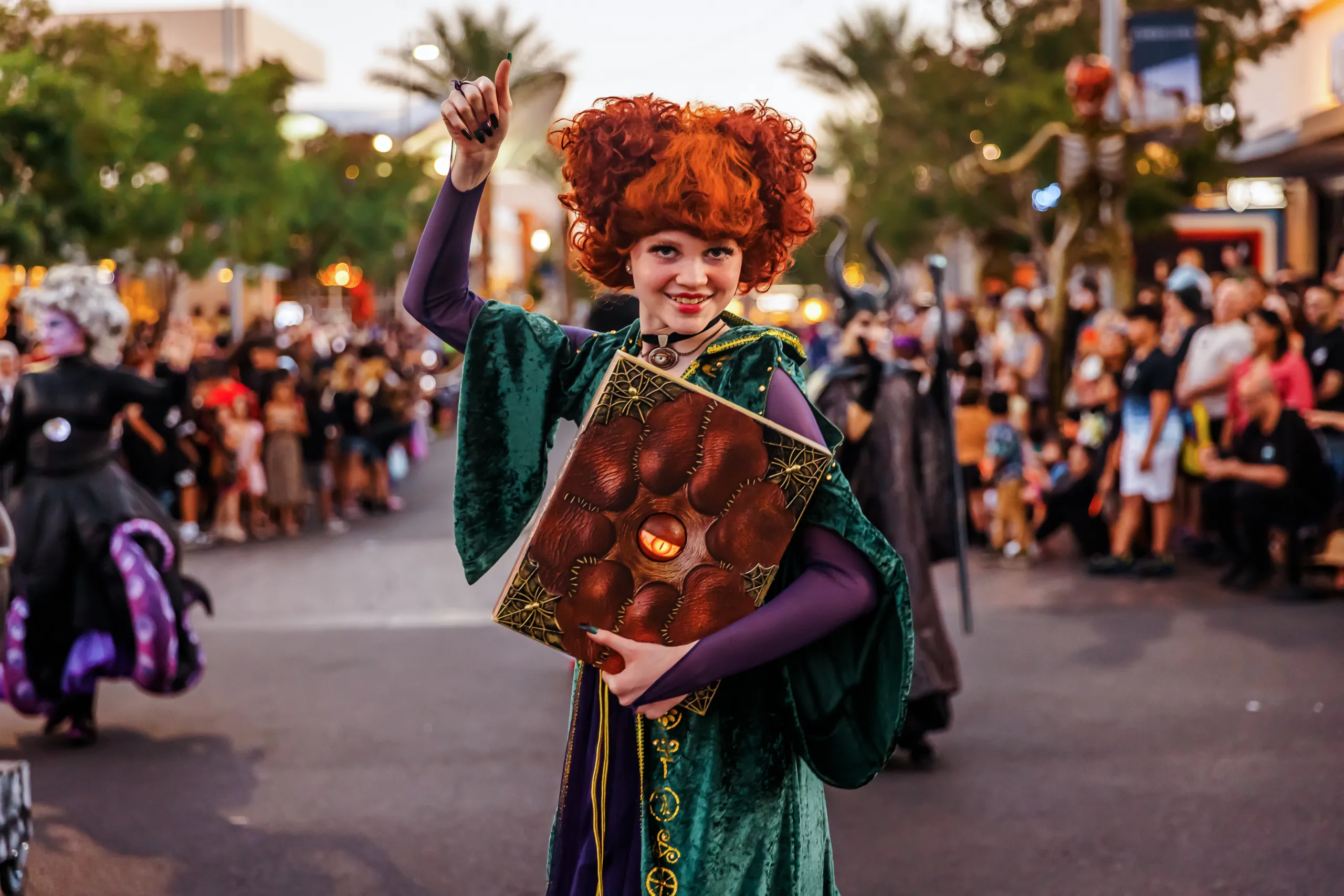 Person in a green witch costume with bright red curly hair holds a large, brown spellbook while smiling at a parade. Blurry crowd and palm trees are visible in the background.