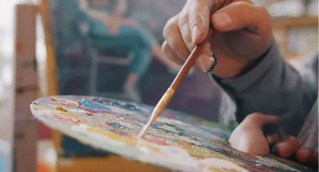 Event Guide A close-up of a hand holding a paintbrush, mixing colors on a painters palette, with a blurred figure seated in the background.