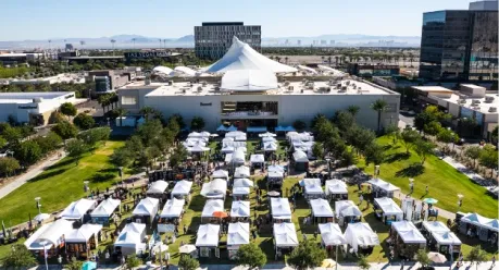 Event Map Aerial view of an outdoor event with rows of white tents on a lawn, people walking between them, and a large modern building in the background under a clear sky.