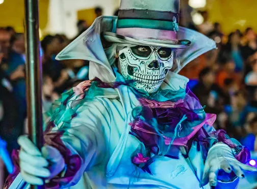 Person in an elaborate Day of the Dead costume with face painted as a skeleton, wearing a decorated top hat and colorful, ornate clothing, posing in a crowd—capturing the vibrant spirit often seen at community events near Summerlin residences.