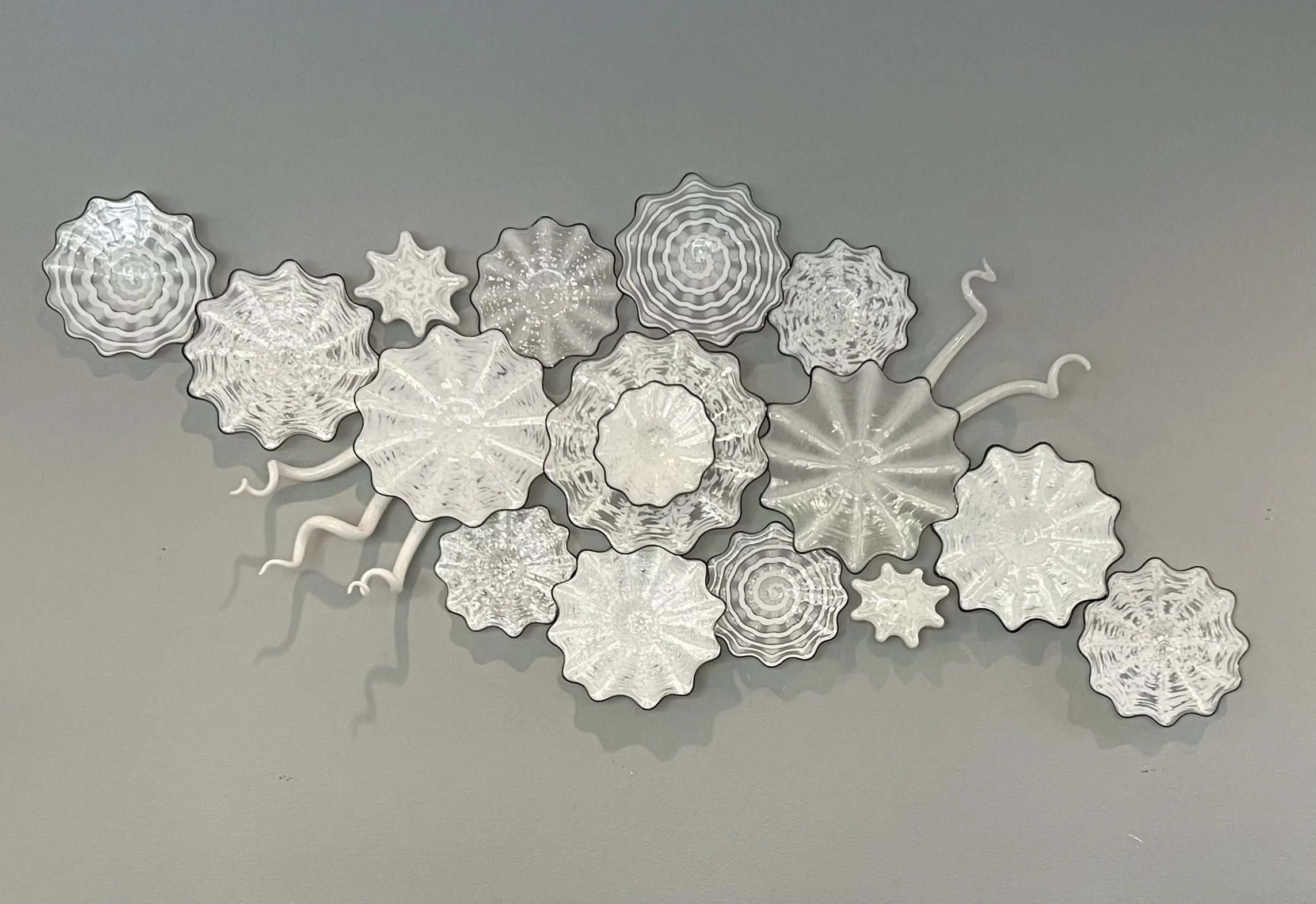 Glass art installation featuring multiple round, textured, translucent white pieces with various patterns, arranged to resemble abstract sea forms or flowers, mounted on a light gray wall. White wavy elements extend outward.