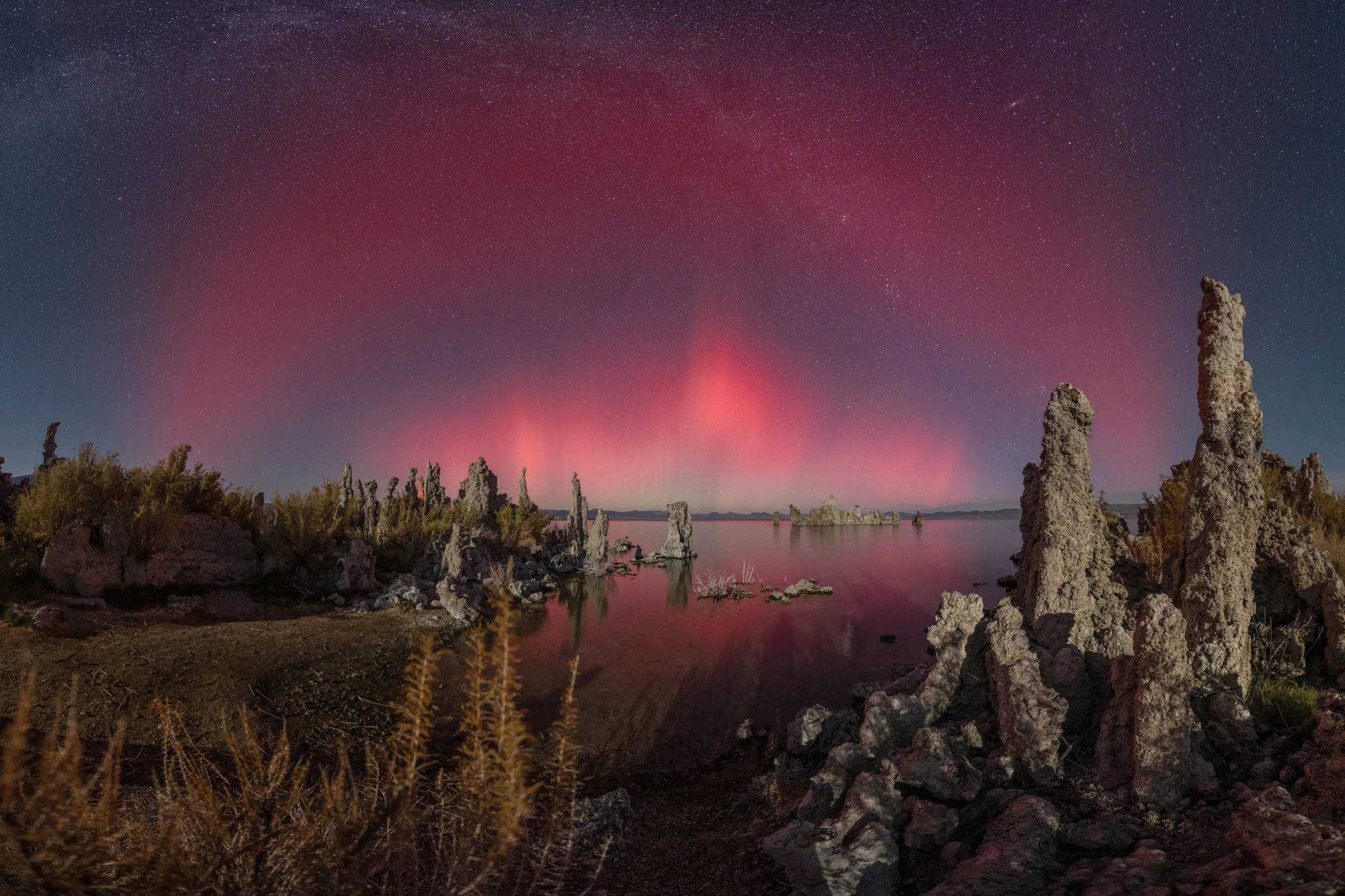 Tall rock formations surround a calm lake under a night sky filled with bright pink and red aurora lights, reflecting beautifully on the water. Sparse vegetation appears in the foreground.
