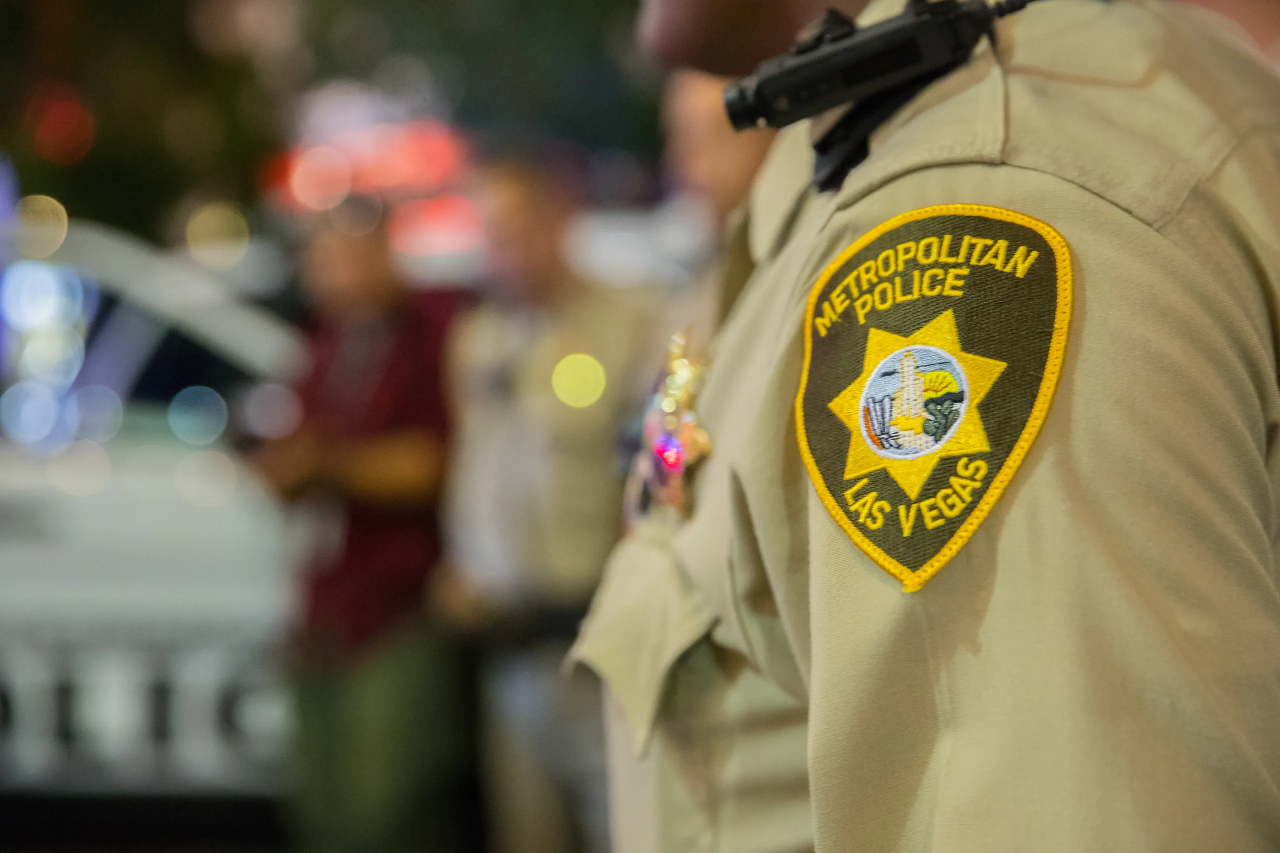 A close-up of a Metropolitan Police Las Vegas officers uniform patch, with a blurred background showing other people and part of a police vehicle.