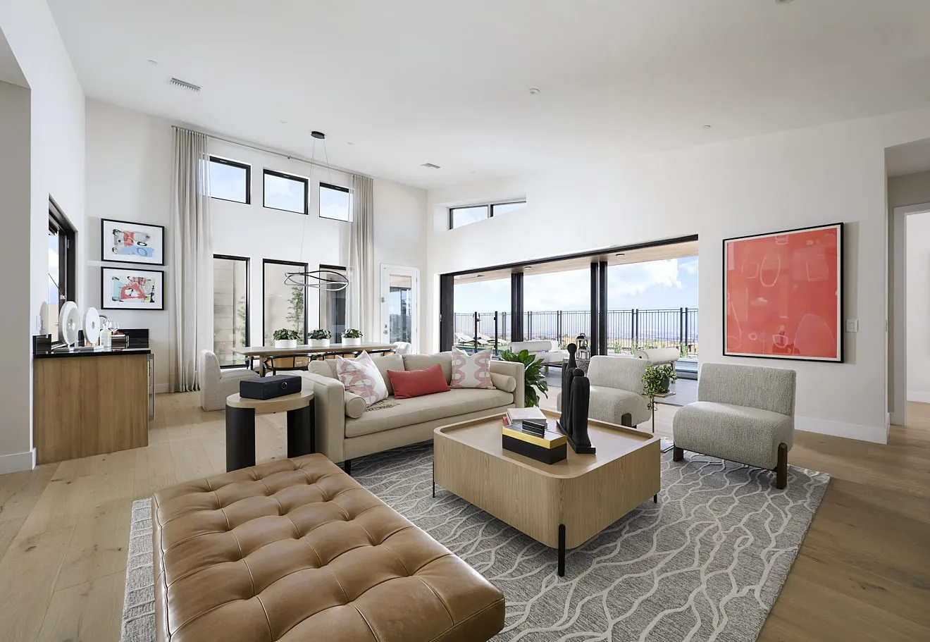 Modern living room with large windows, neutral furniture, a tan leather bench, two armchairs, a light rug, and abstract art on white walls. Sliding doors open to a balcony with a view.