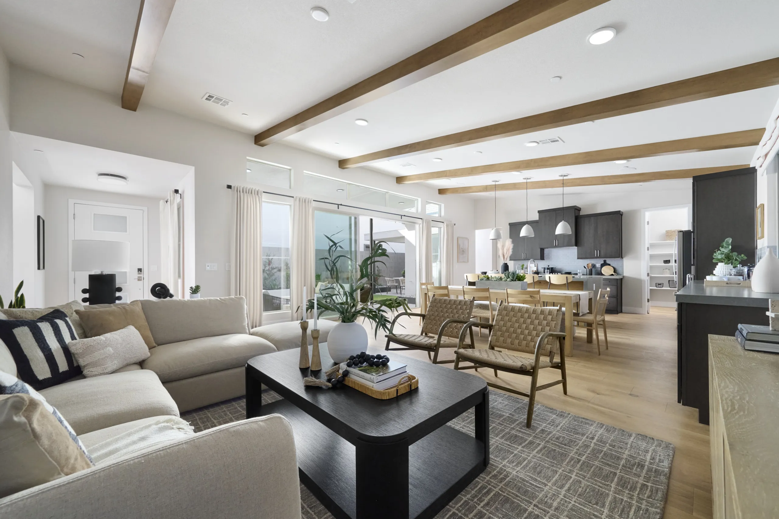 Modern open-concept living room with beige sofa, wooden chairs, black coffee table, dining area, and kitchen. Large windows let in natural light; exposed ceiling beams and plants add warmth to the space.