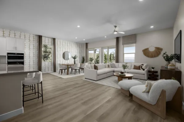 Spacious modern living room with light wood flooring, white sectional sofa, accent chairs, dining area, large windows, and neutral decor. Kitchen counter with barstools is visible on the left.