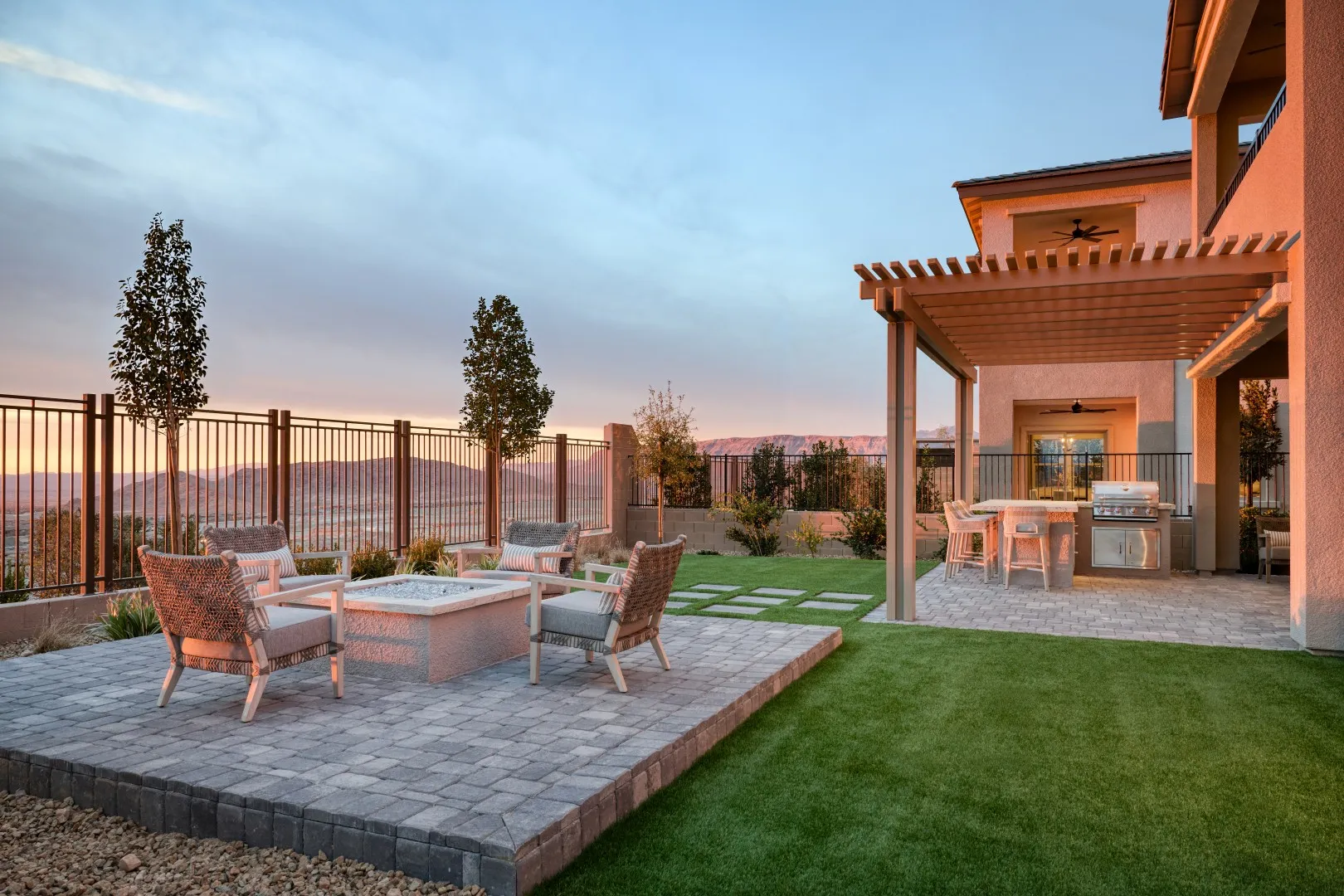 Modern backyard with a patio seating area around a firepit, green lawn, pergola-covered dining space, barbecue grill, and mountain views at sunset.