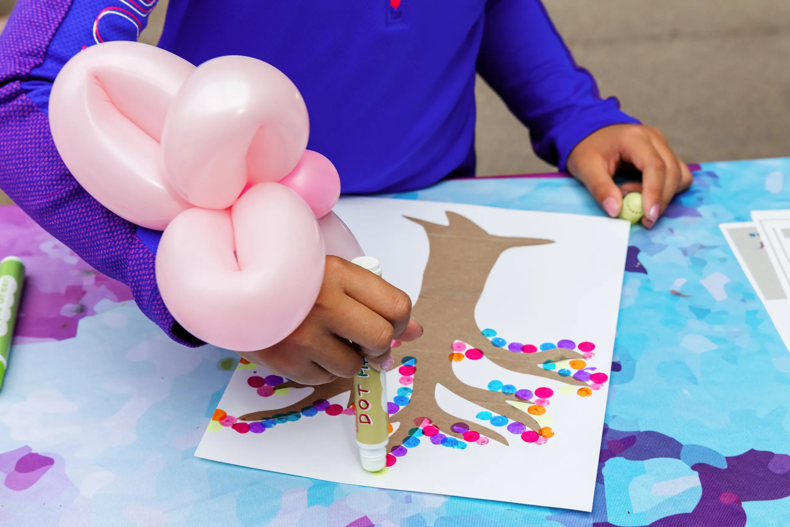 A child wearing a pink balloon bracelet decorates a paper tree with colorful dot markers at a table covered with a blue patterned cloth.