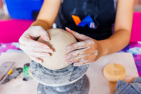 A person with flour-covered hands shapes a round ball of clay on a pottery wheel, wearing a dark apron. A sponge and cloth sit on the table nearby.