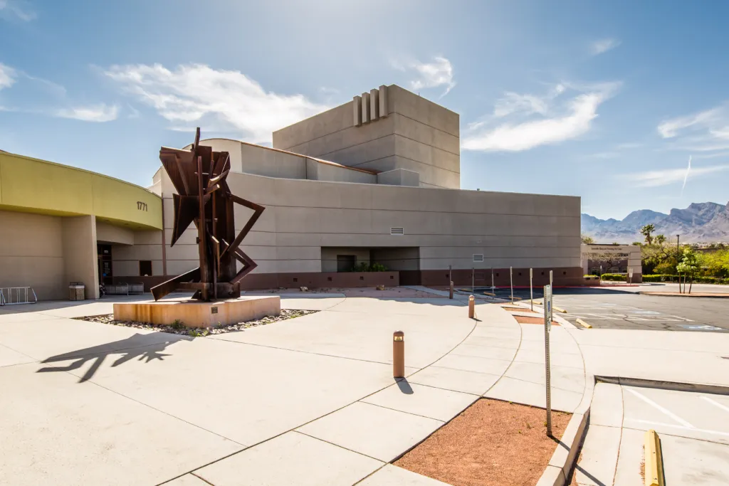 Modern beige building with geometric architecture, a large abstract metal sculpture in front, and mountains visible in the background under a sunny sky.