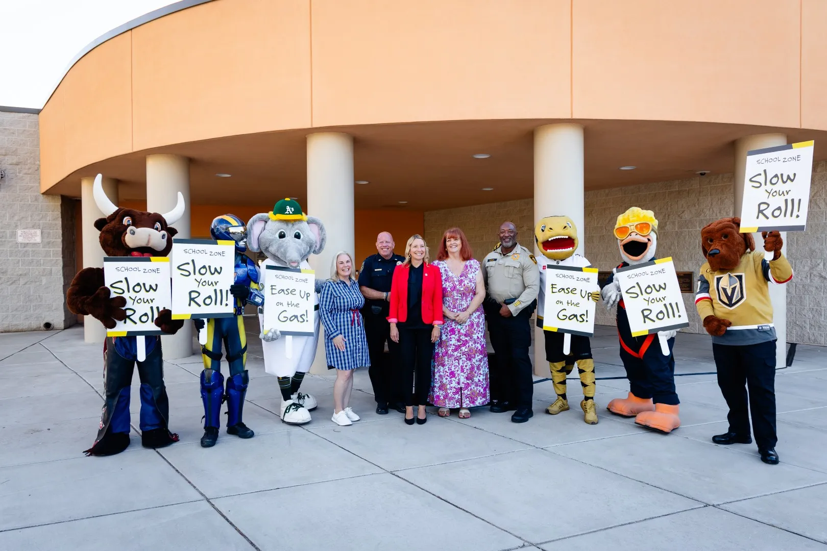 A group of people and mascots stand together outside a building. The mascots hold signs that say Slow Your Roll! and Ease Up on the Gas! promoting safe driving in school zones.