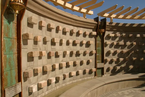 Curved memorial wall with engraved plaques and square stones, under a pergola with sunlight casting shadows. There is a bronze lamp and a bench along the walls base.