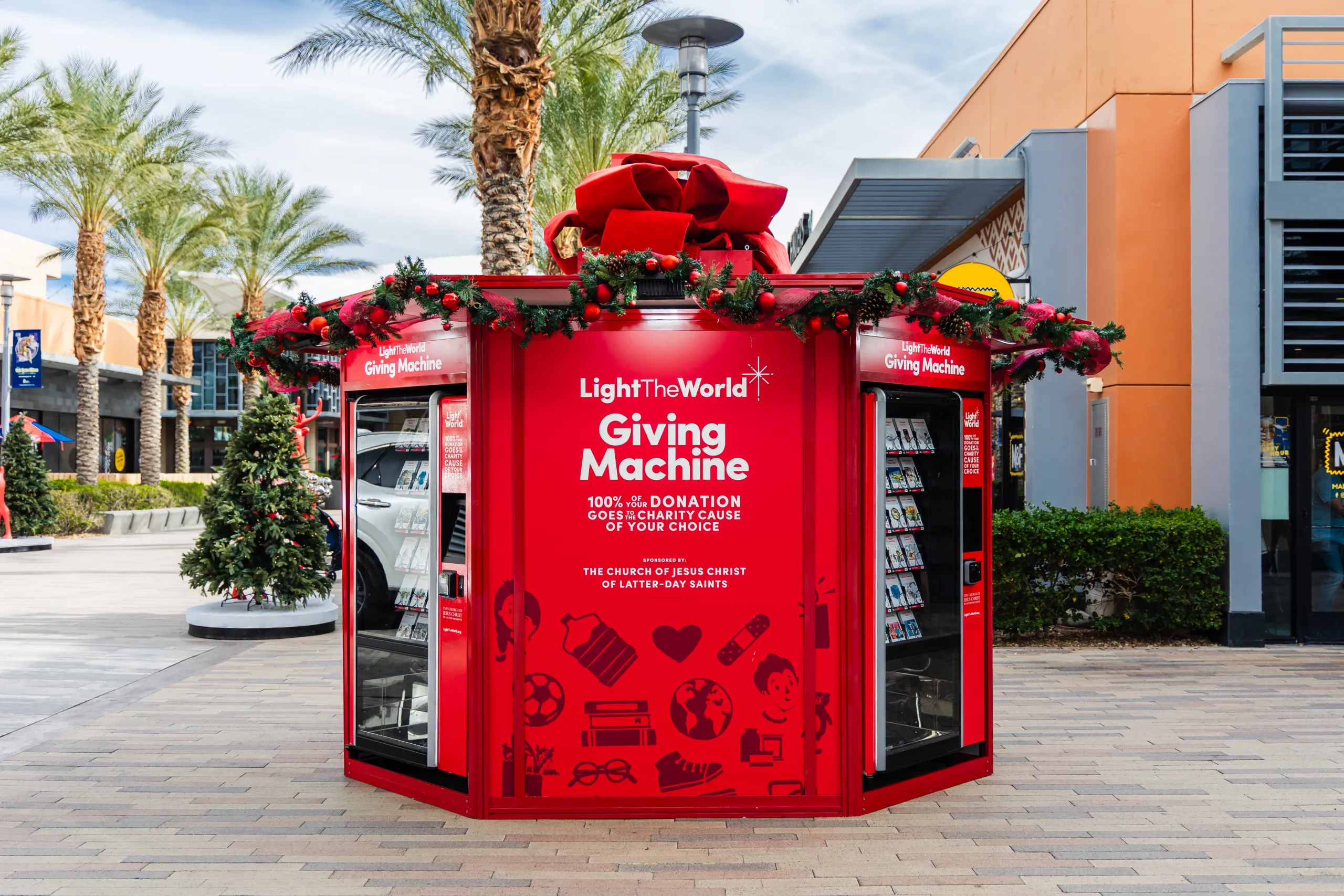 A bright red LightTheWorld Giving Machine kiosk decorated with a large bow and garland, standing in an outdoor shopping plaza.