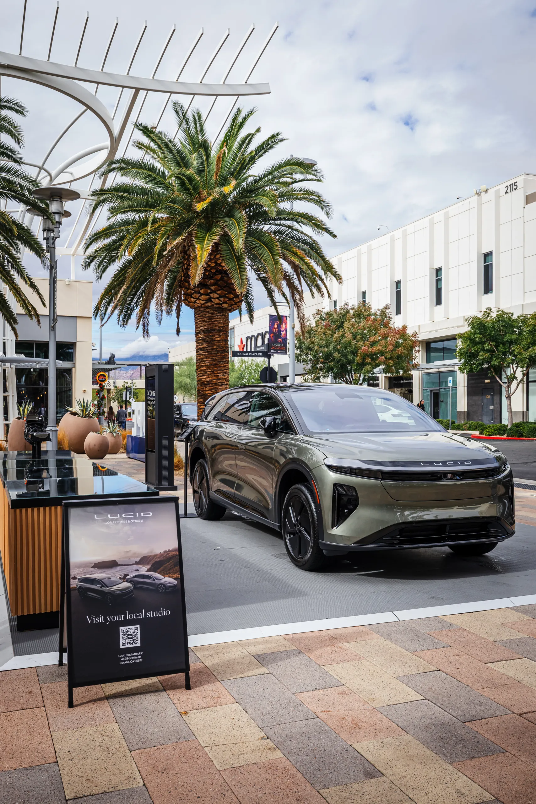 Olive green Lucid Gravity SUV parked in an outdoor shopping plaza next to a branded display sign with a QR code.