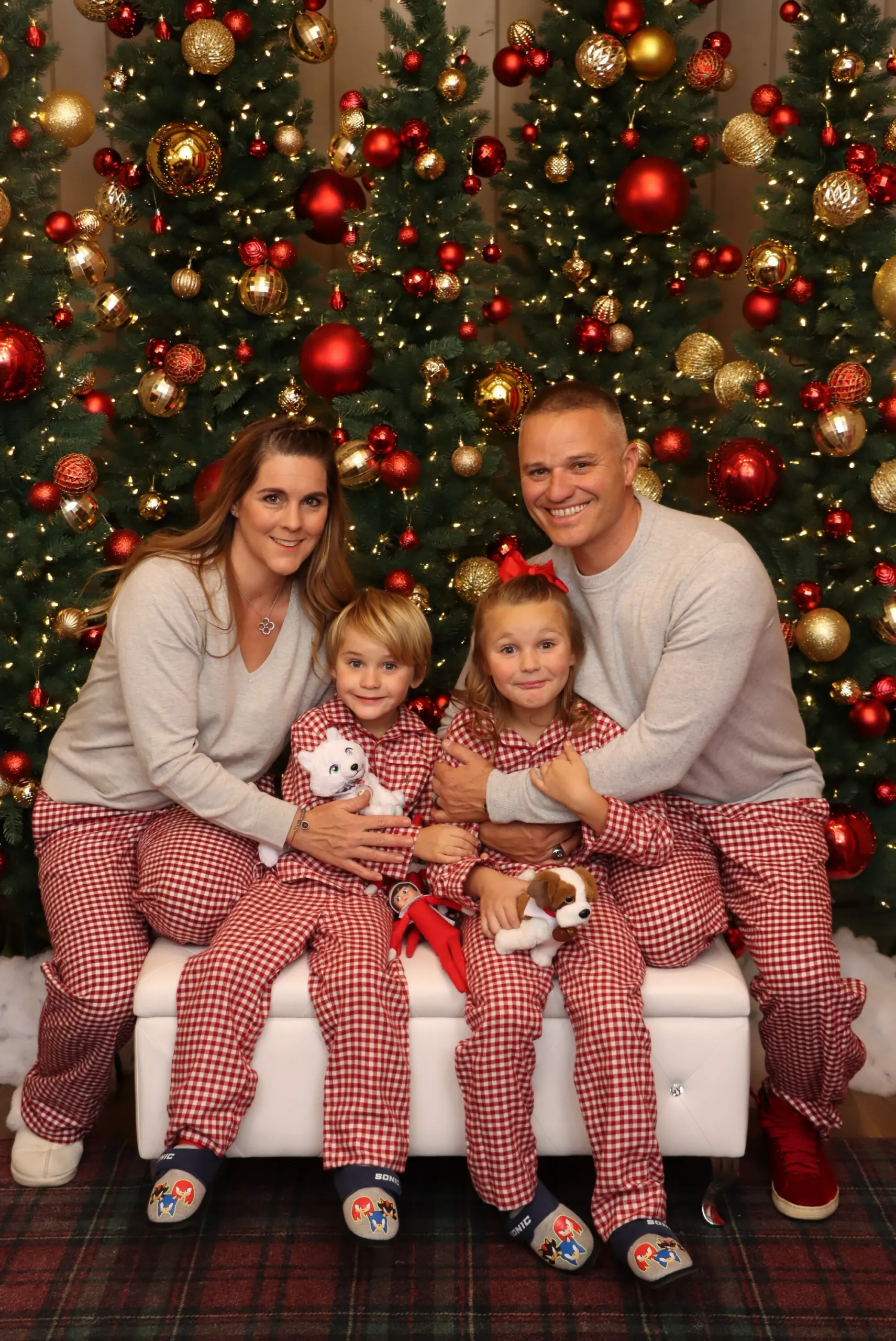Holiday Family Photos A family of four wearing matching red and white plaid pajamas sits on a bench in front of two decorated Christmas trees.