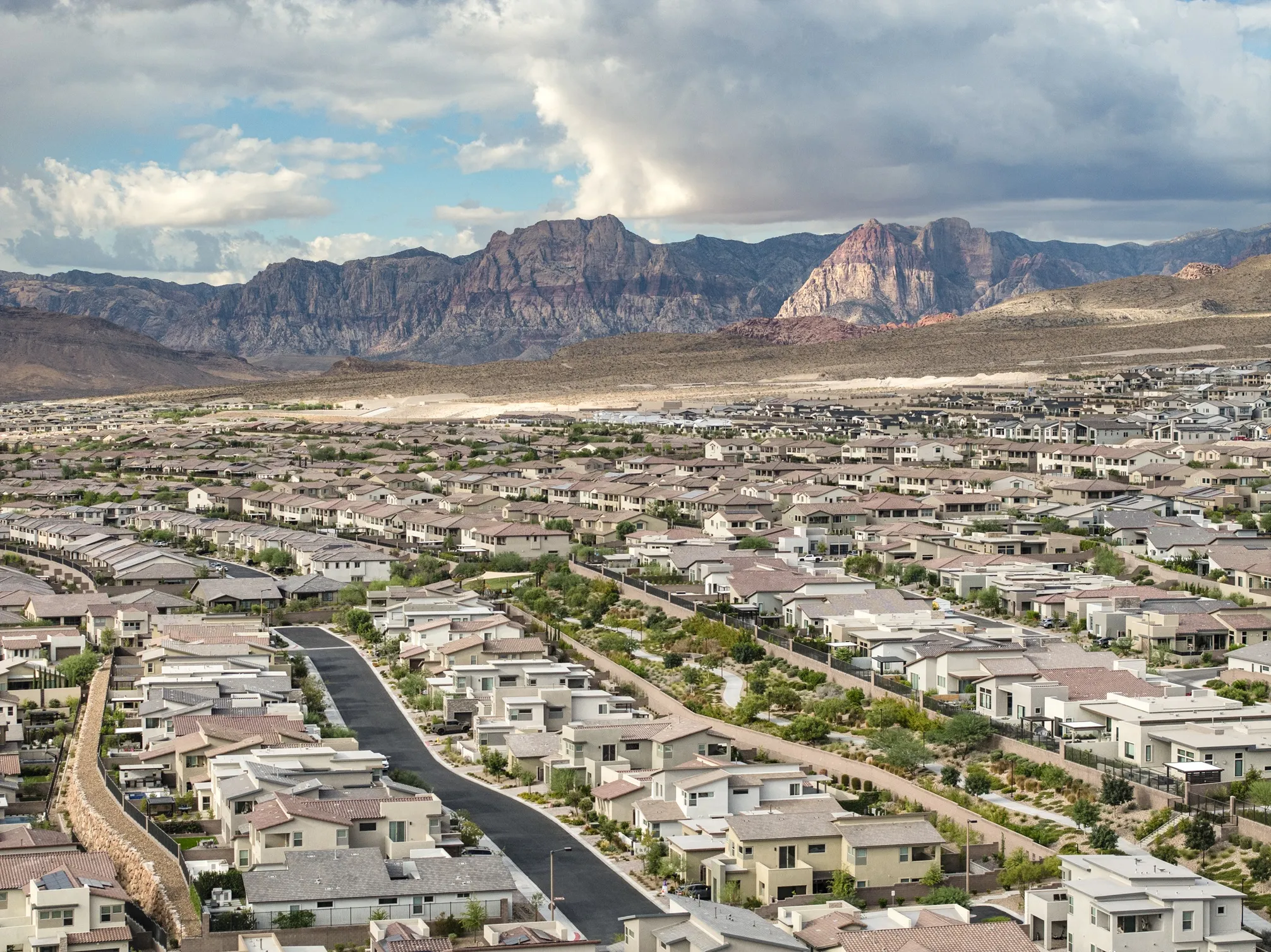 Aerial view of a sprawling residential neighborhood of suburban homes with a backdrop of large, rugged mountains under a cloudy sky.