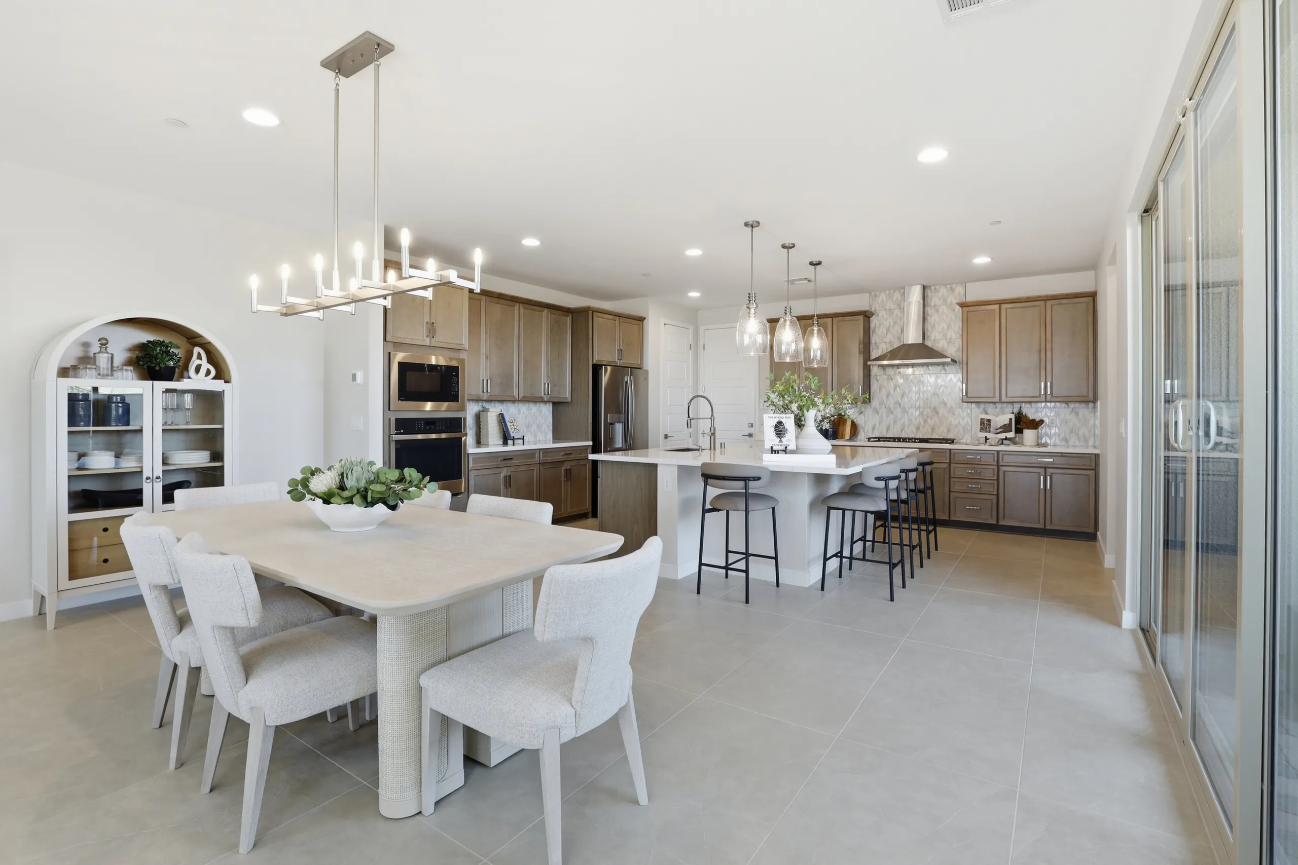 A bright, open-concept kitchen and dining area featuring a light-toned dining table with six upholstered chairs, a large kitchen island with bar seating, and wooden cabinetry.