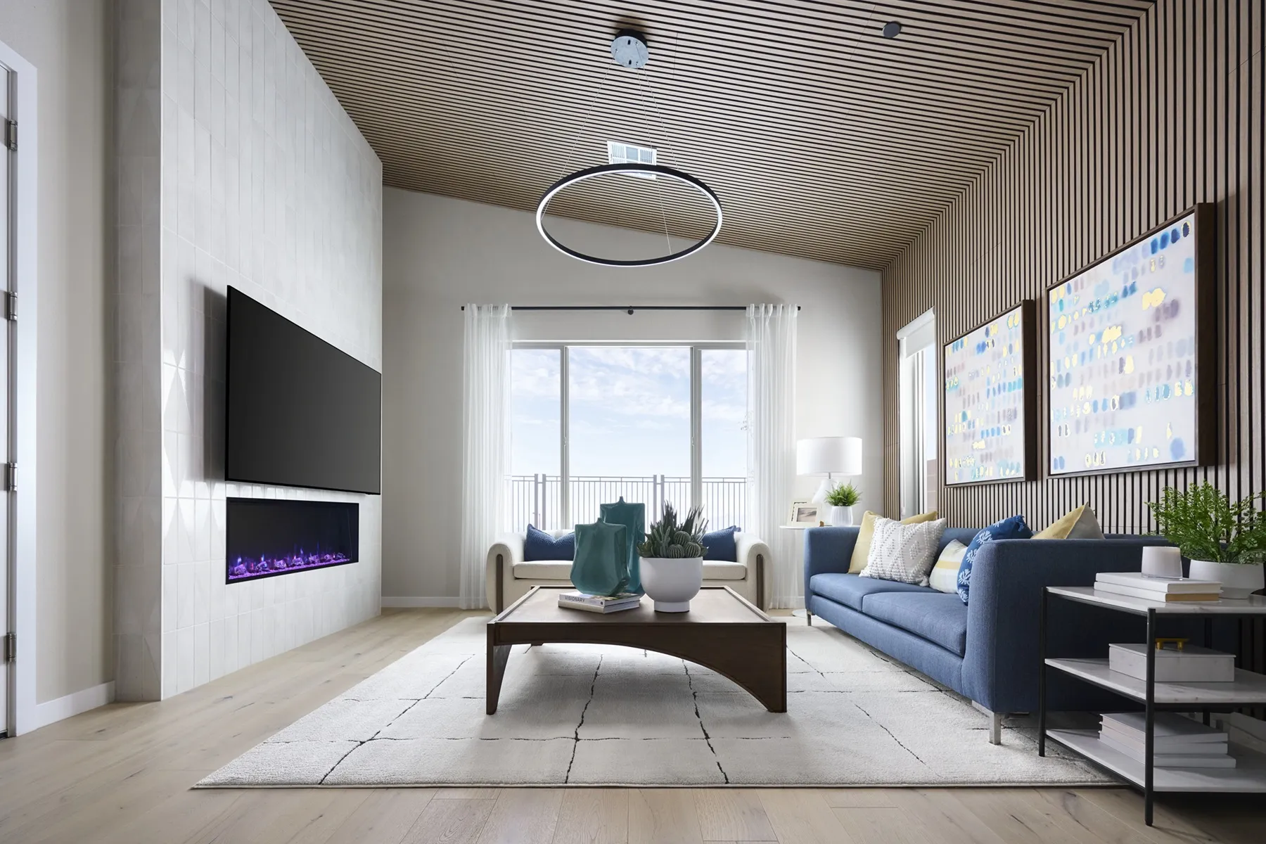 Modern living room with a blue sofa, a linear fireplace below a wall-mounted television, a wooden slat ceiling, and a large circular pendant light.