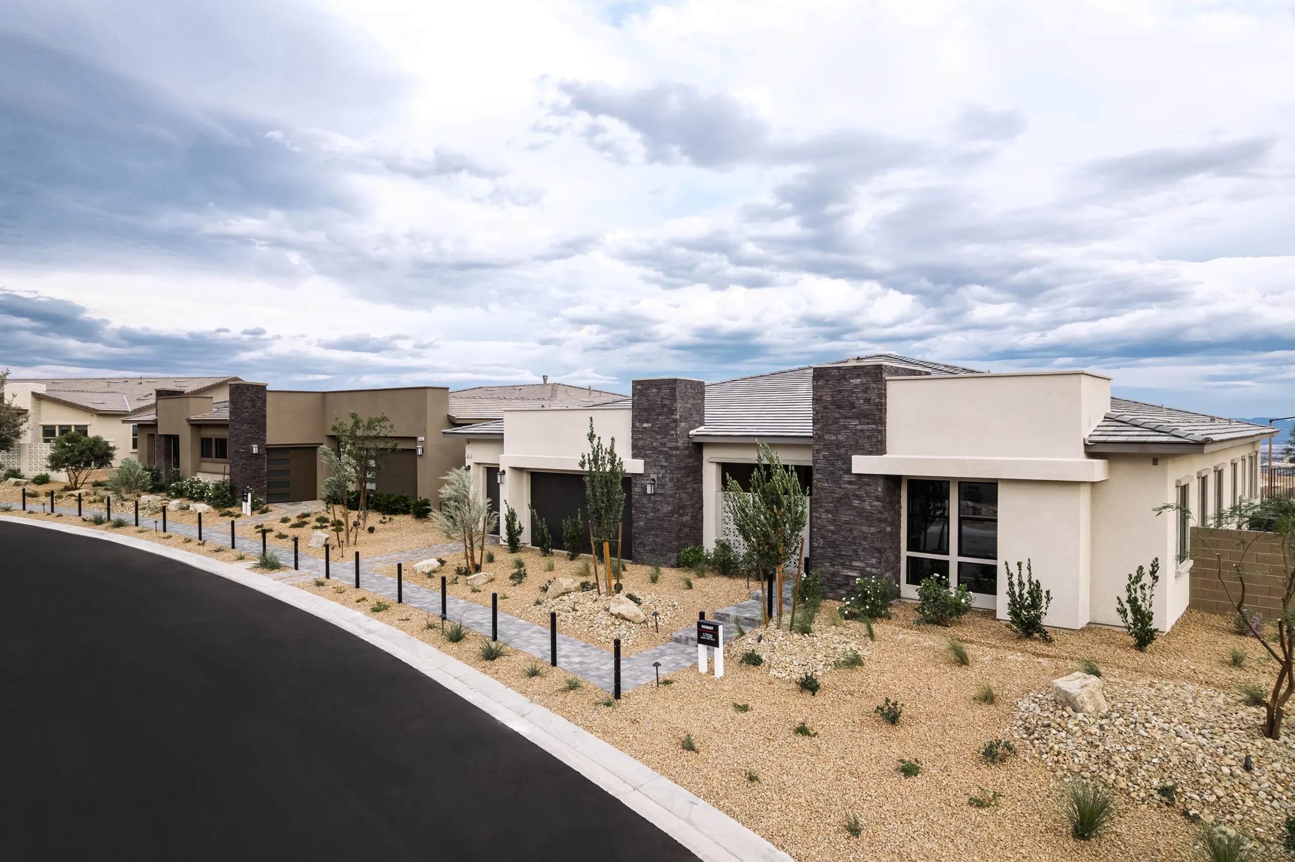 A row of modern single-story homes with stucco and stone facades and desert landscaping along a curved asphalt street.