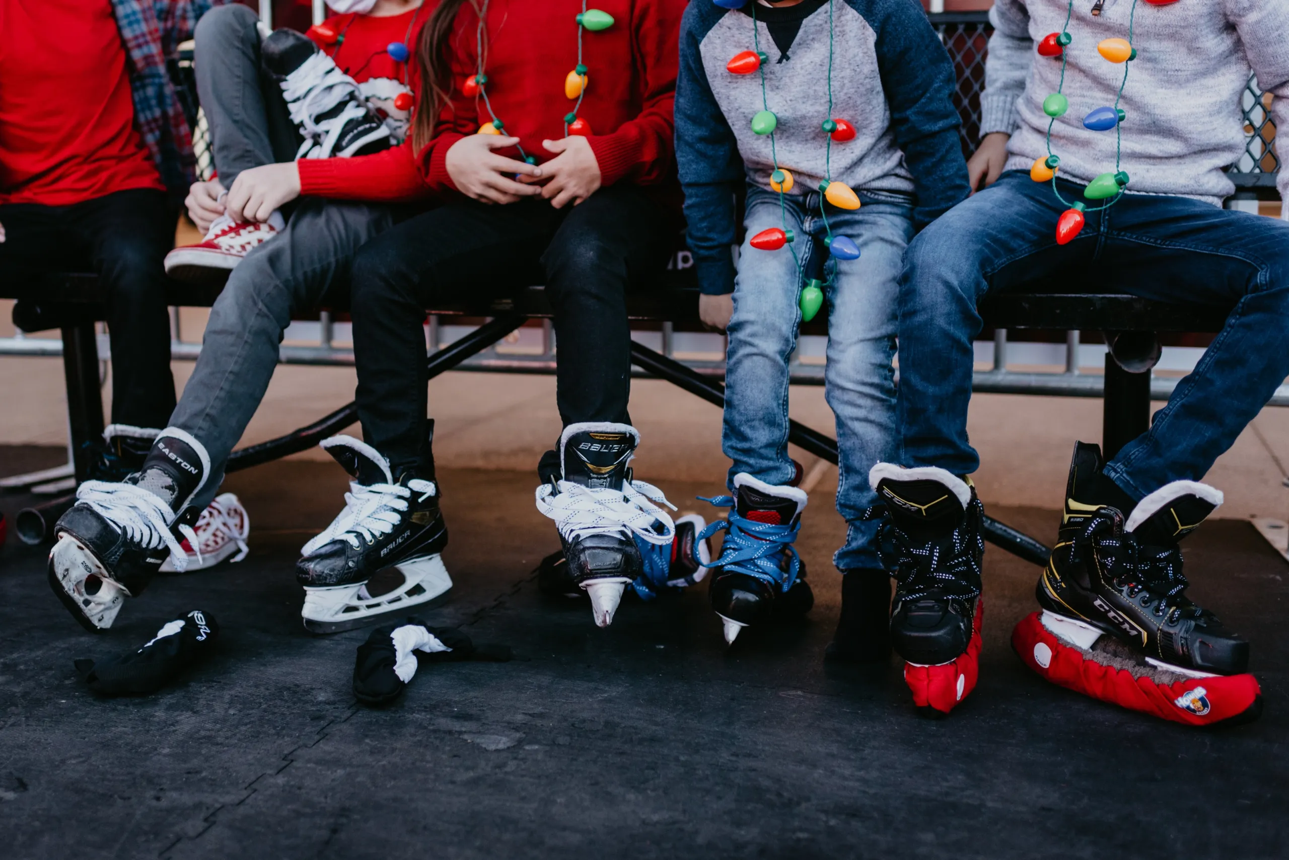 A group of people wearing sweaters adorned with string lights sit on a bench, wearing ice skates on their feet.