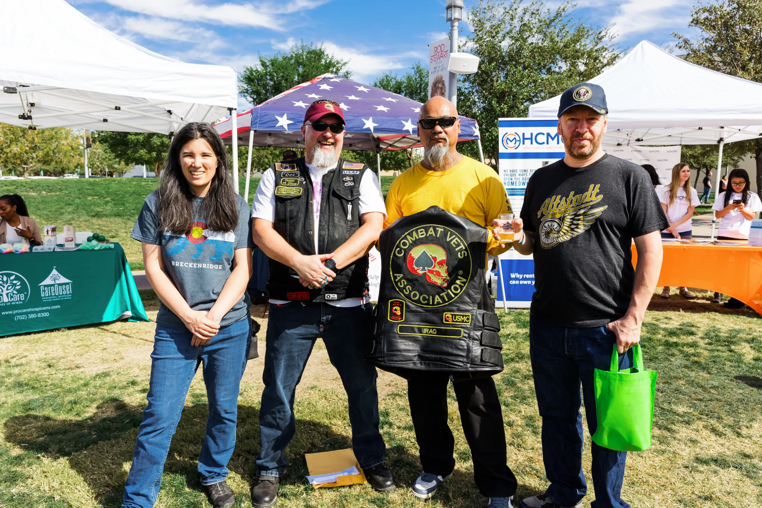 Four people stand together outdoors at a community event, with one man in the center holding up a black leather Combat Vets Association vest.