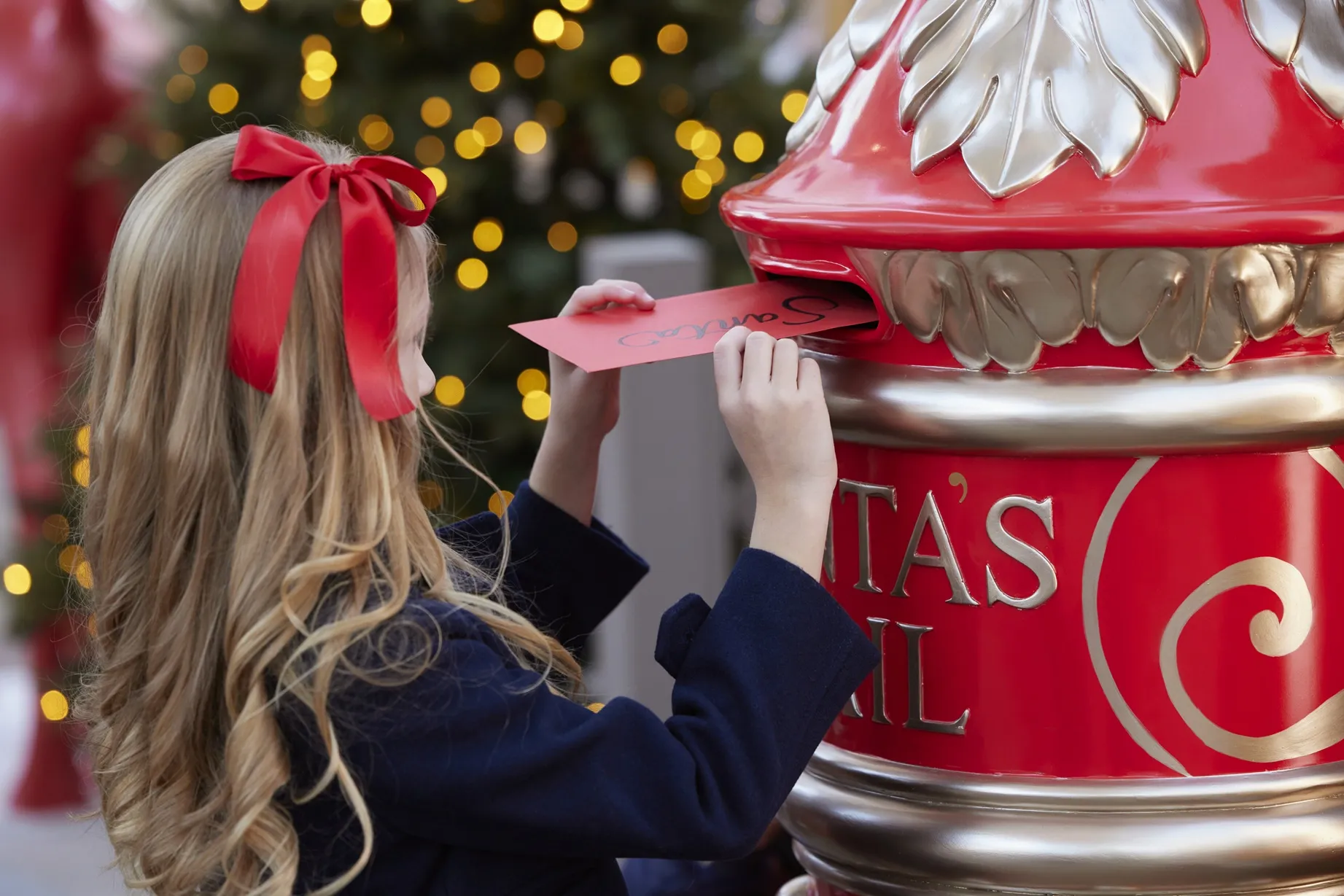 A young girl with a red bow in her hair drops a letter addressed to Santa into a red and gold mailbox.