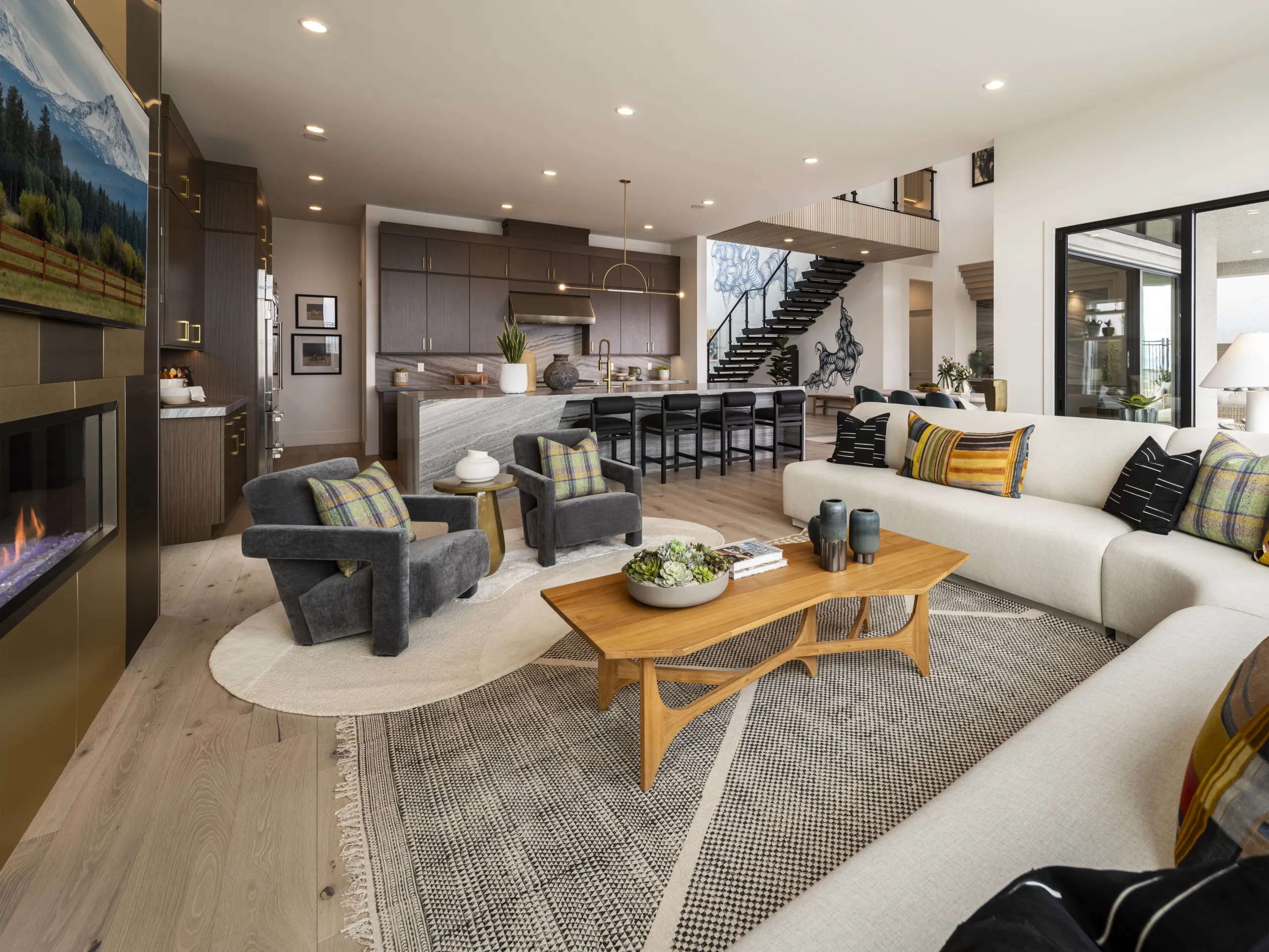 Open-plan living room with a cream sectional, wooden coffee table, two gray armchairs, and a modern kitchen with dark cabinetry in the background.